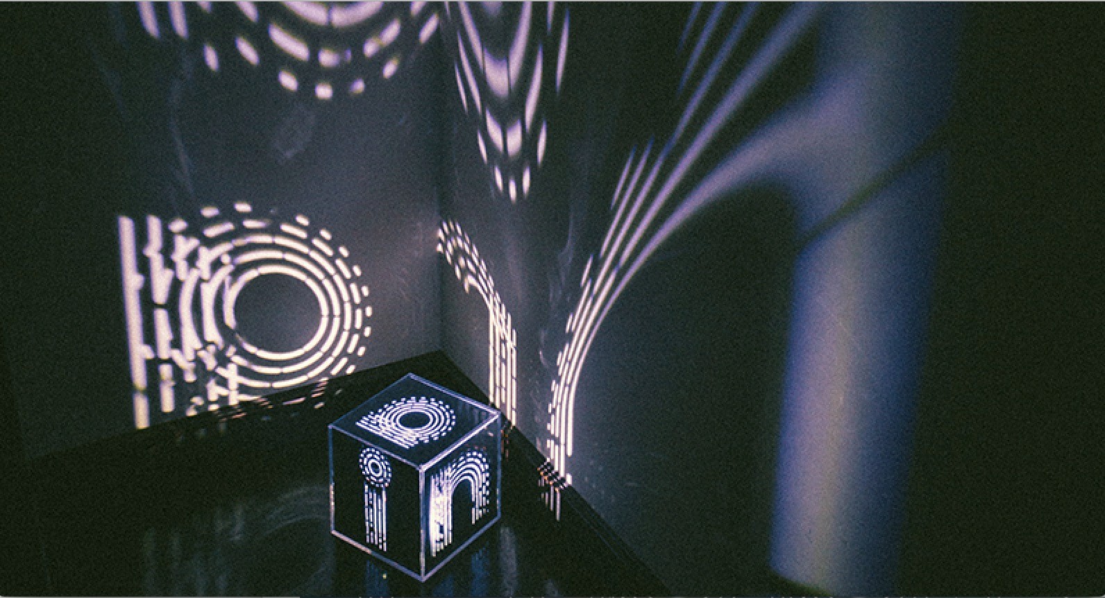 A photo of a cube that has on each side a letter. The light emitted from the cube illuminates the walls near by with the letters from each side of the cube.