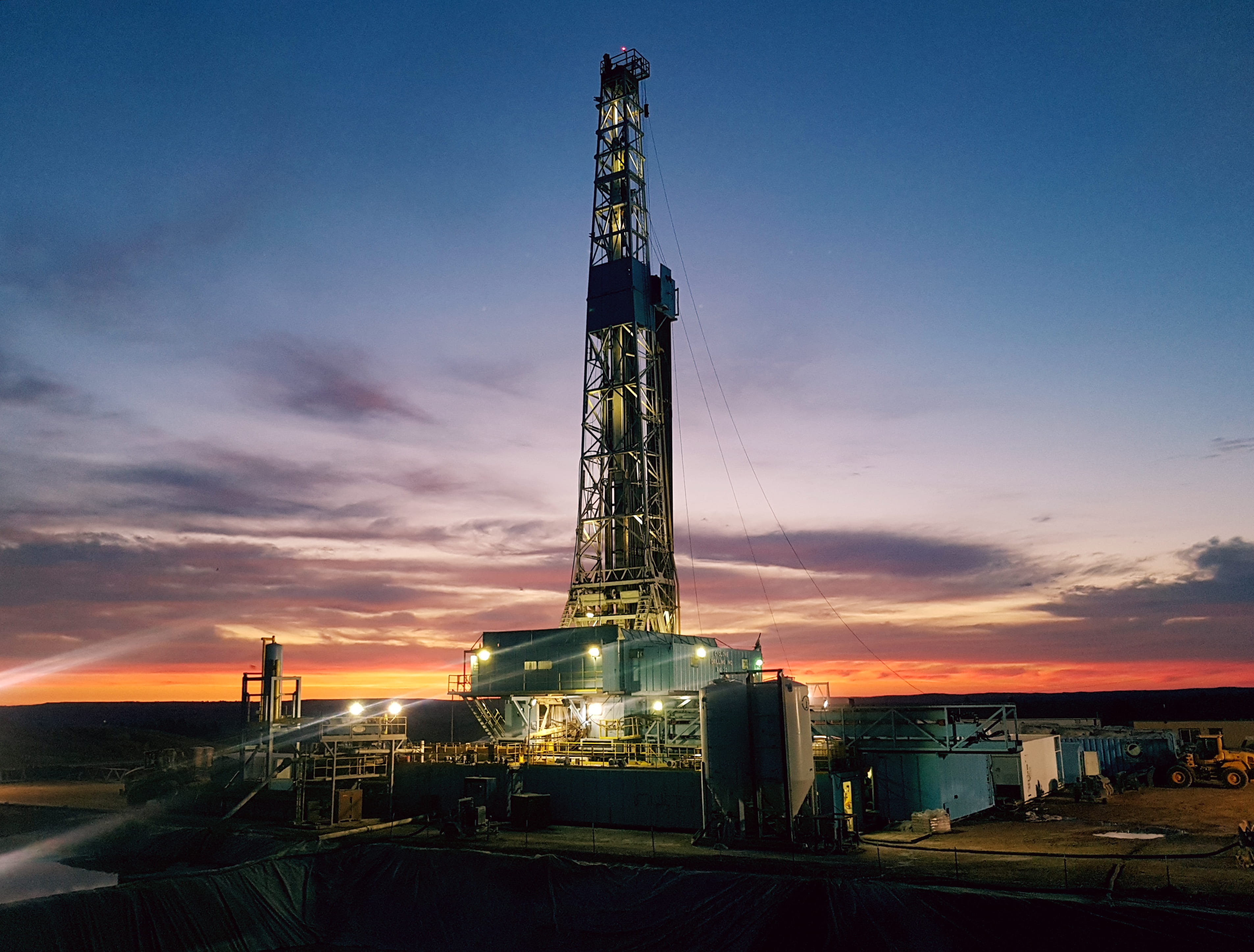 An illuminated land drilling rig operating in a flat desert landscape during a vibrant orange and purple sunset.