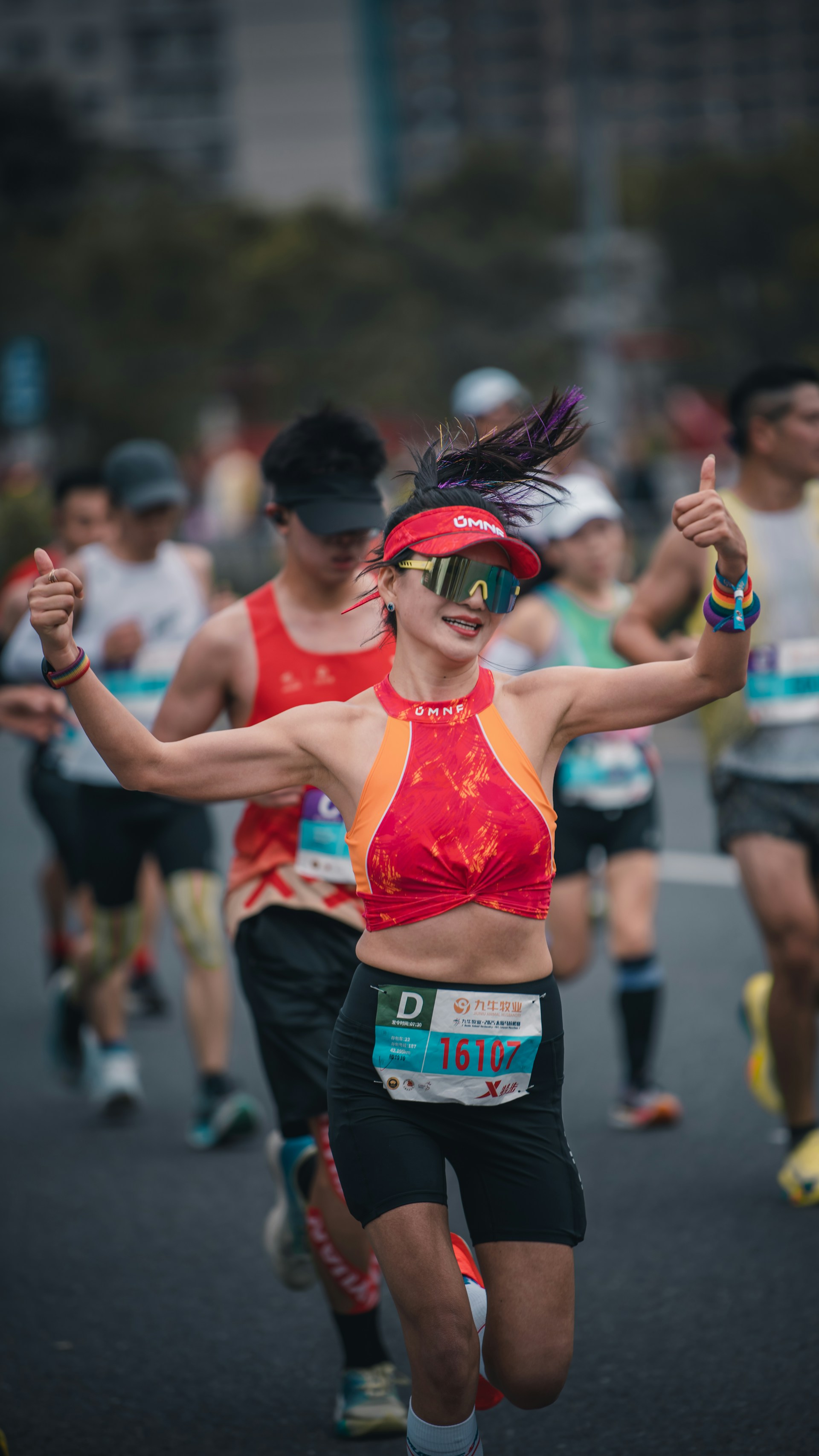 A smiling female marathon runner in a red top and sunglasses giving two thumbs up to the camera during a race.