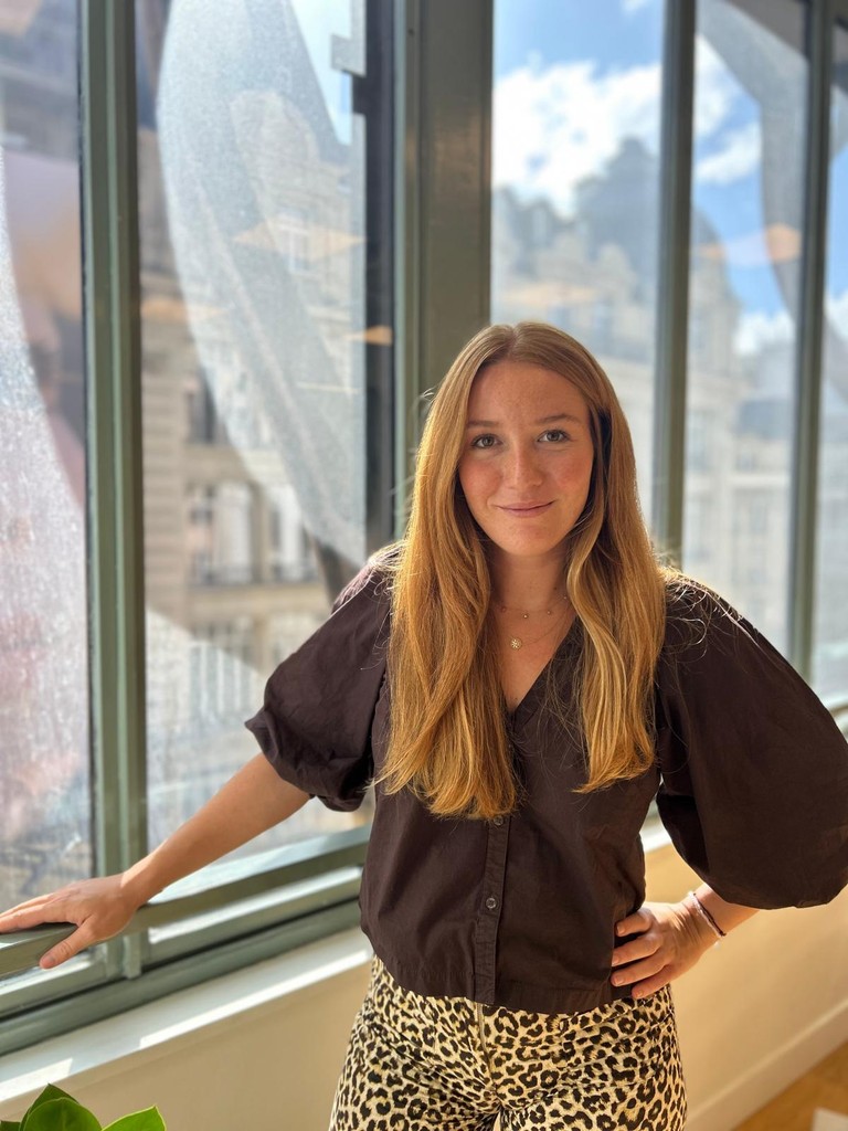 Young person with curly hair wearing a rust-colored blazer and black turtleneck, looking confidently at the camera against a neutral background.