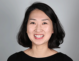 The image shows a woman with short, dark hair against a plain, light gray background. She is wearing a black top and smiling warmly at the camera. Her facial features are clearly visible, including her eyes, nose, and mouth. The image is a headshot, capturing her face and upper body prominently. There are no additional objects or text in the image.