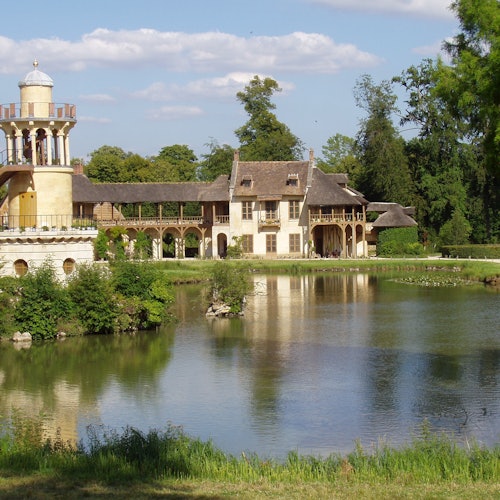 A picturesque building with a tower beside a calm pond, surrounded by greenery and trees under a partly cloudy sky.