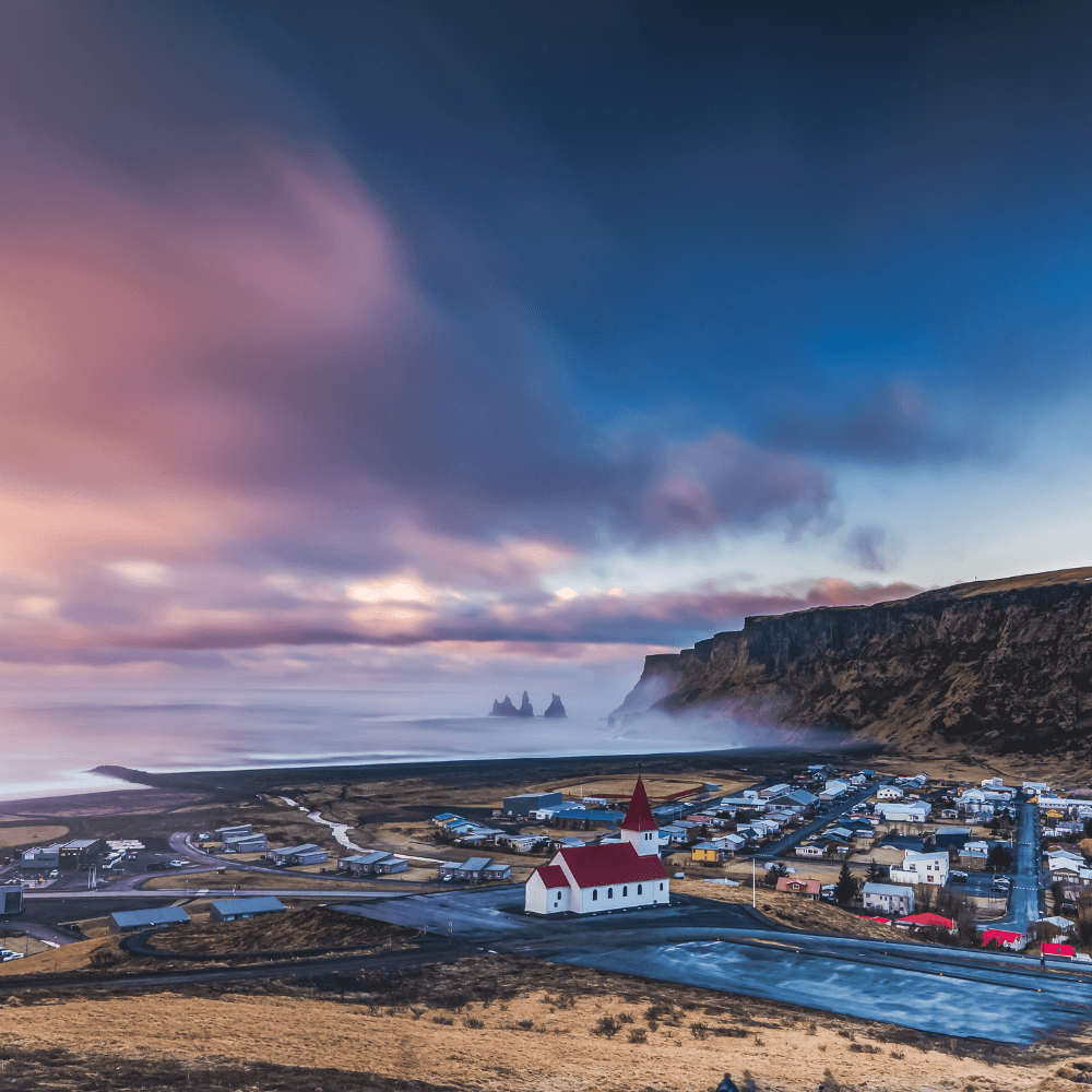 Panoramic image of Vík í Mýrdal in South Iceland showing the red church, black sand beach, Reynisdrangar sea stacks, coastal cliffs, and village landscape at sunset.