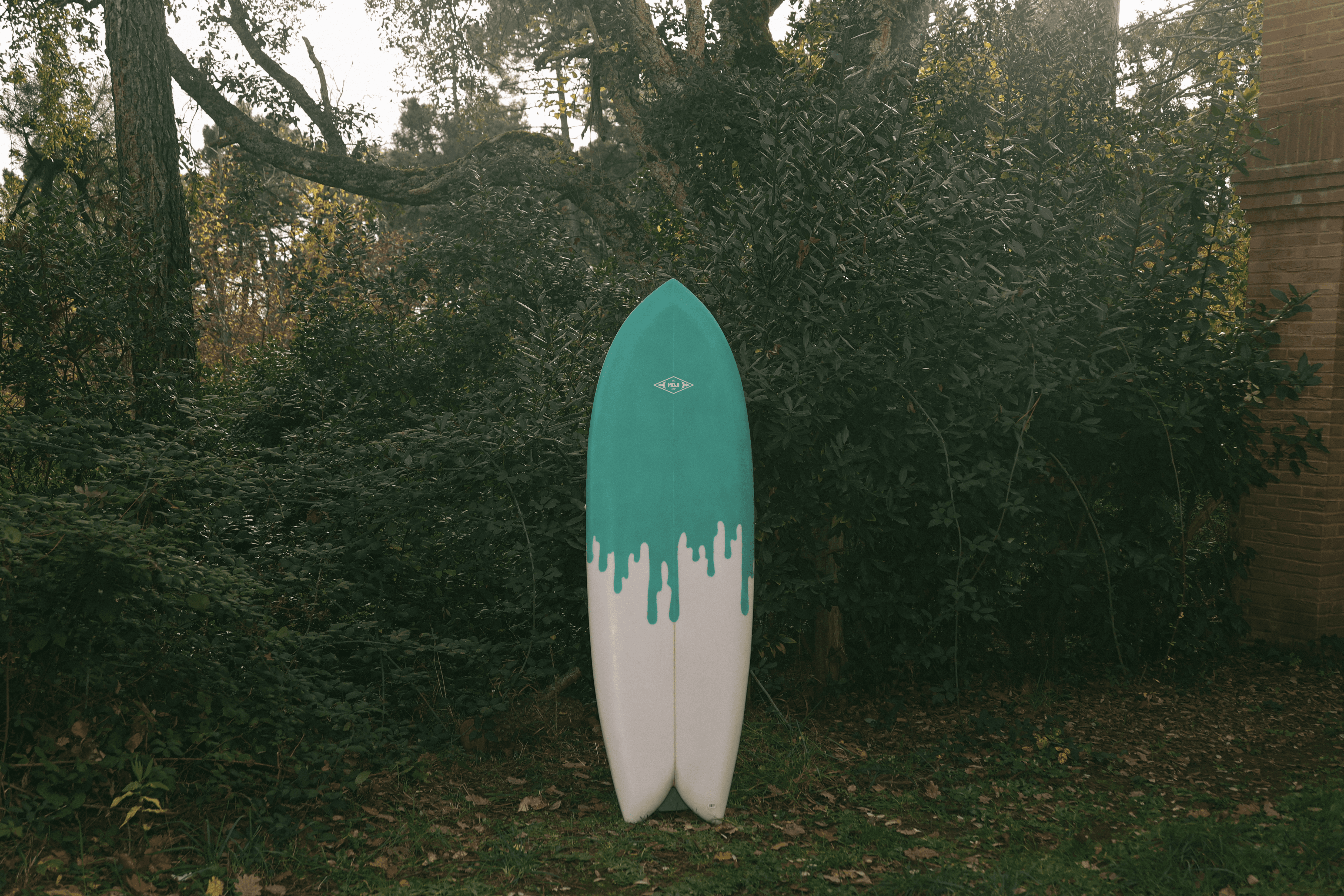 Tavola da surf modello Lady Finger bianca e verde acqua, con disegno a gocciolamento e configurazione a due pinne, esposta verticalmente in un'area boscosa.