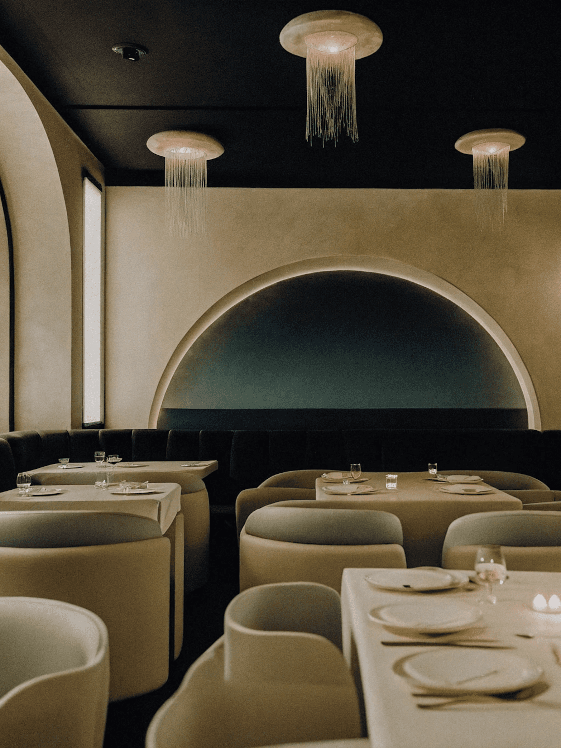 Modern restaurant interior with soft tones, arched walls, jellyfish-like pendant lights, and elegant table settings for a cozy dining vibe.