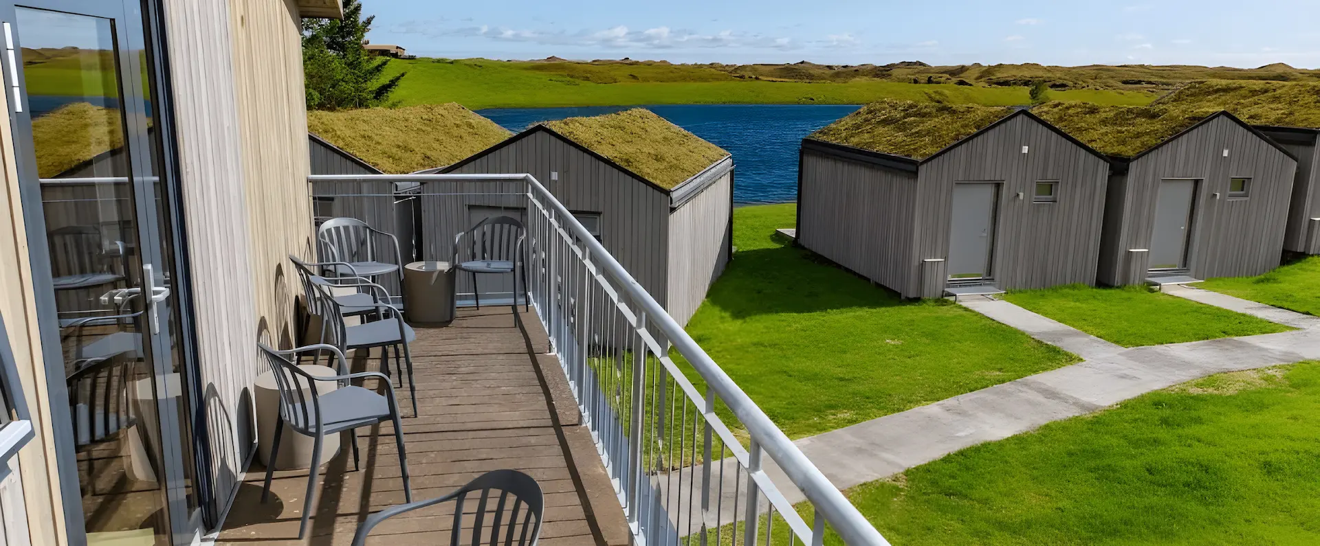 A long rows of modern Nordic cabins with traditional moss-covered grass roofs at Magma Hotel, set against rolling green fields and a blue sky.