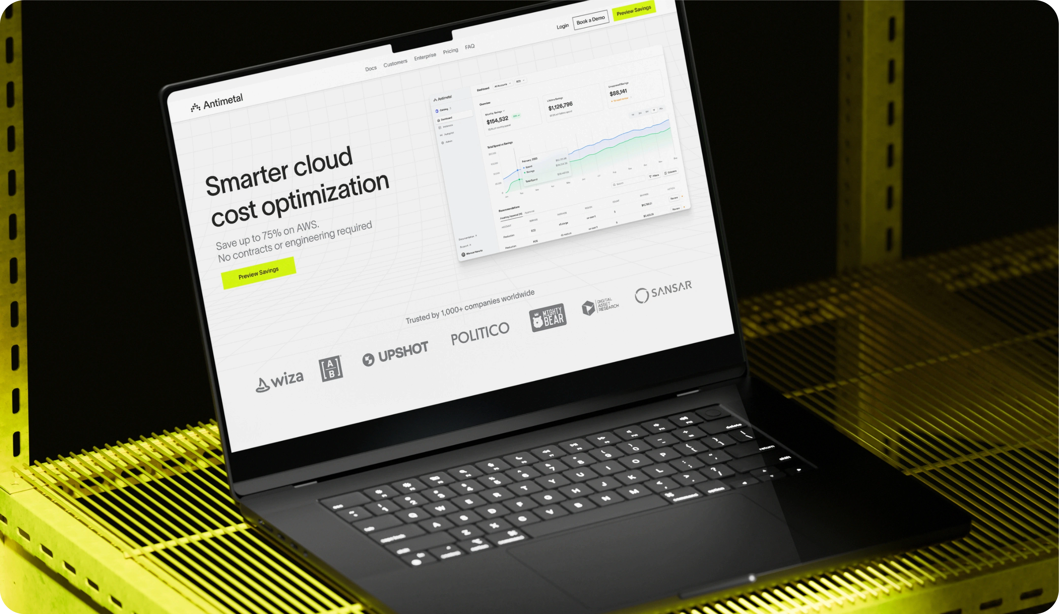 Black laptop displaying Antimetal's website on a yellow wireframe surface. The screen highlights 'Smarter cloud cost optimization' with the claim of saving up to 75% on AWS. Trusted by companies like Wiza, Upshot, Politico, and others, the website invites users to preview savings.