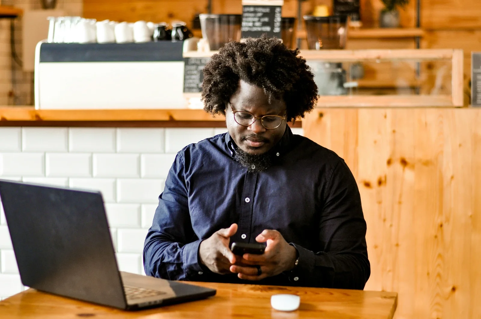 Man in navy shirt using smartphone at wooden table with laptop in modern cafe setting.