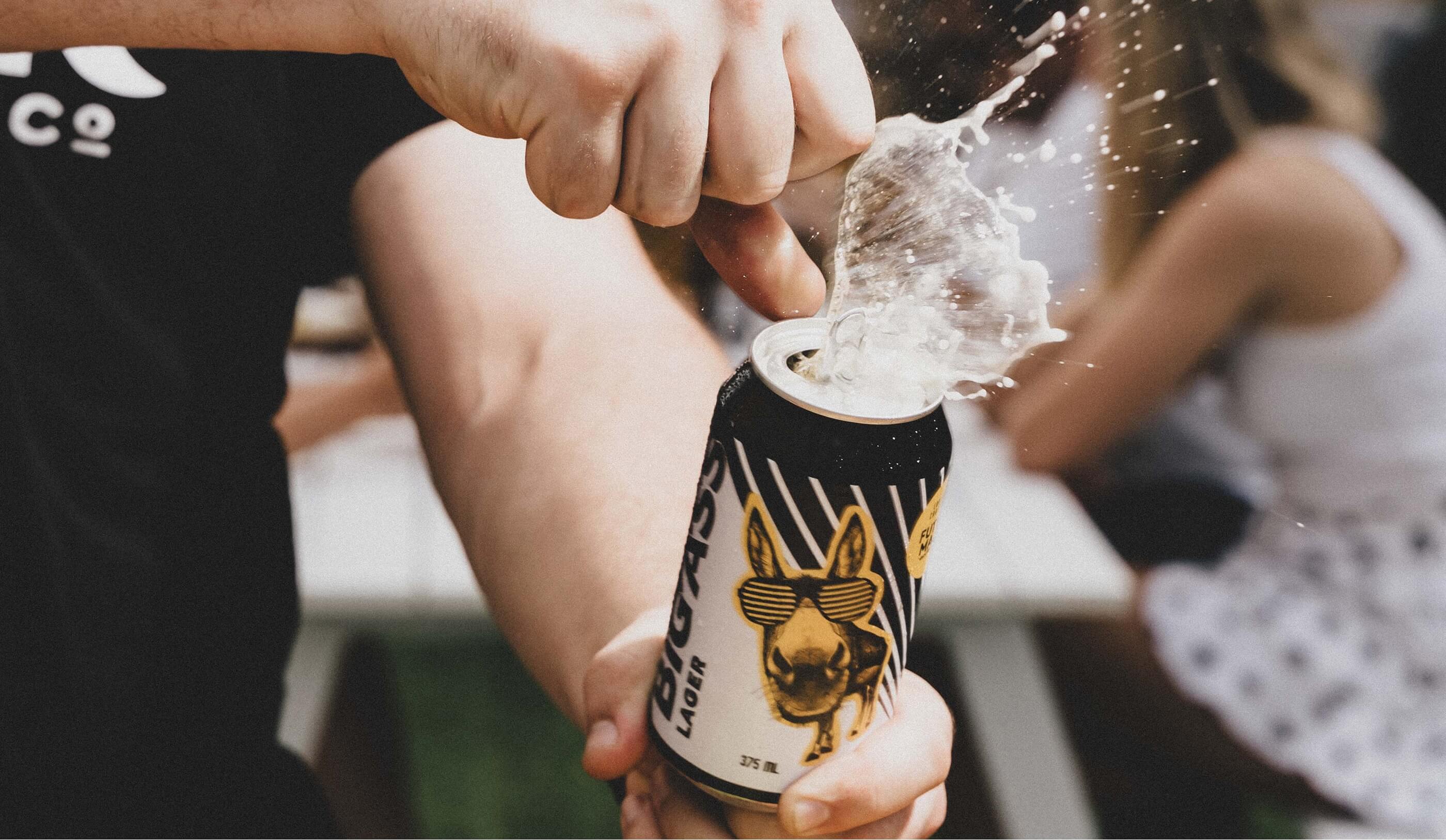 Close-up of a can of Big Ass Lager being opened, with beer foam splashing out, at an outdoor social gathering.