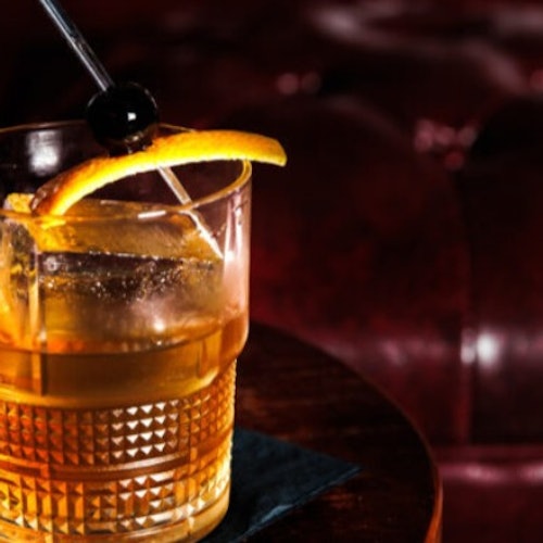 Un vaso de whisky con un cubo de hielo, adornado con una rodaja de naranja y una cereza en un palillo de cóctel, sobre una mesa con un fondo oscuro.