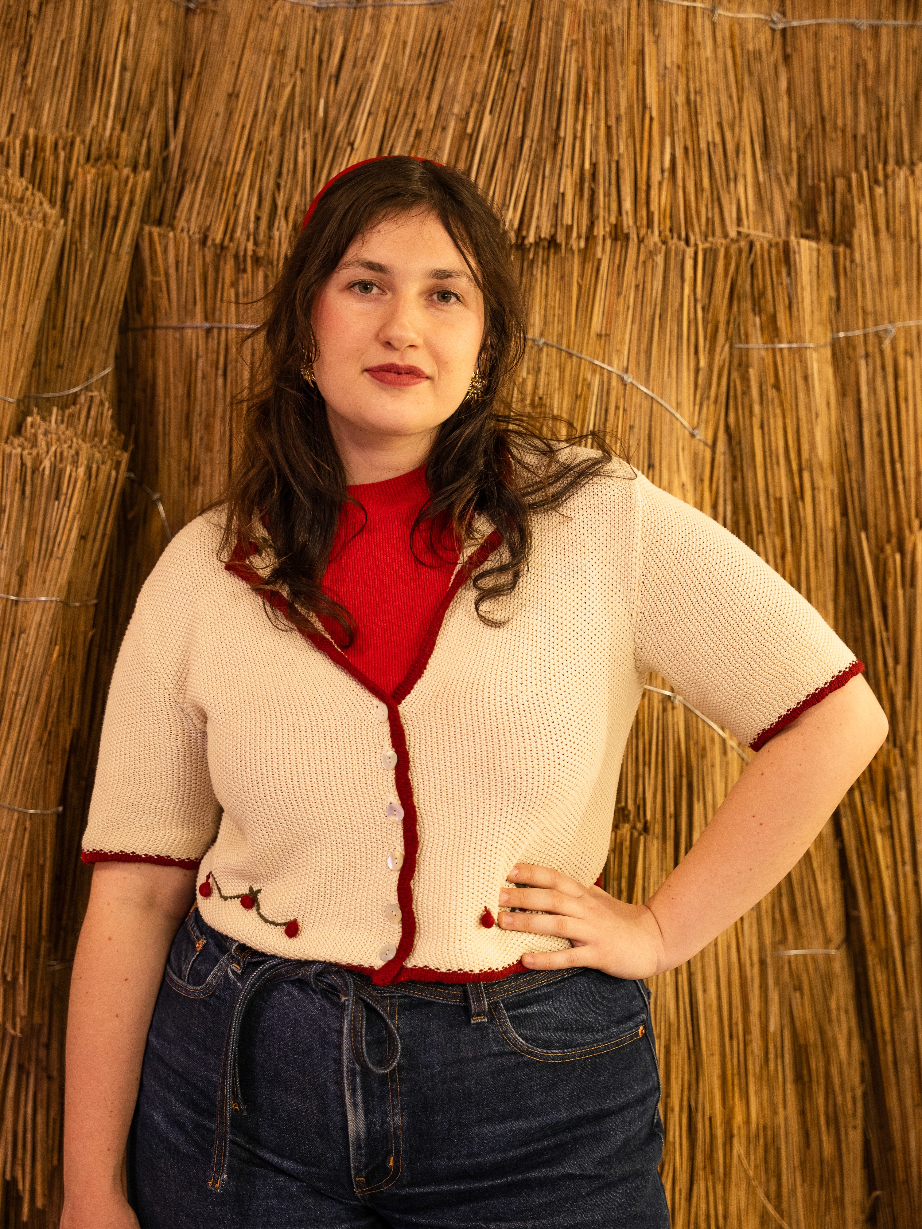 Portrait d’une jeune femme aux cheveux bruns, vêtue d’un gilet crème bordé de rouge sur un haut rouge et d’un jean, posant devant une installation de fibres végétales tressées.