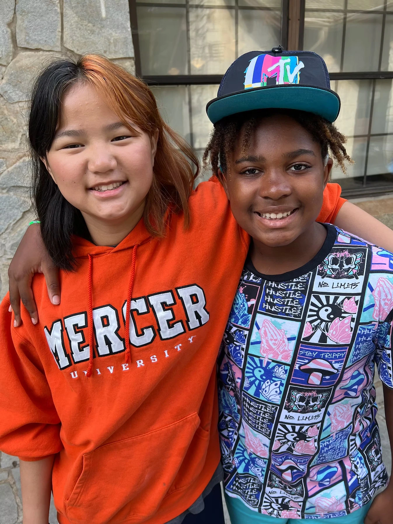 Two smiling teenagers, a girl in an orange Mercer University hoodie and a boy in a colorful shirt and hat, pose with arms around each other.
