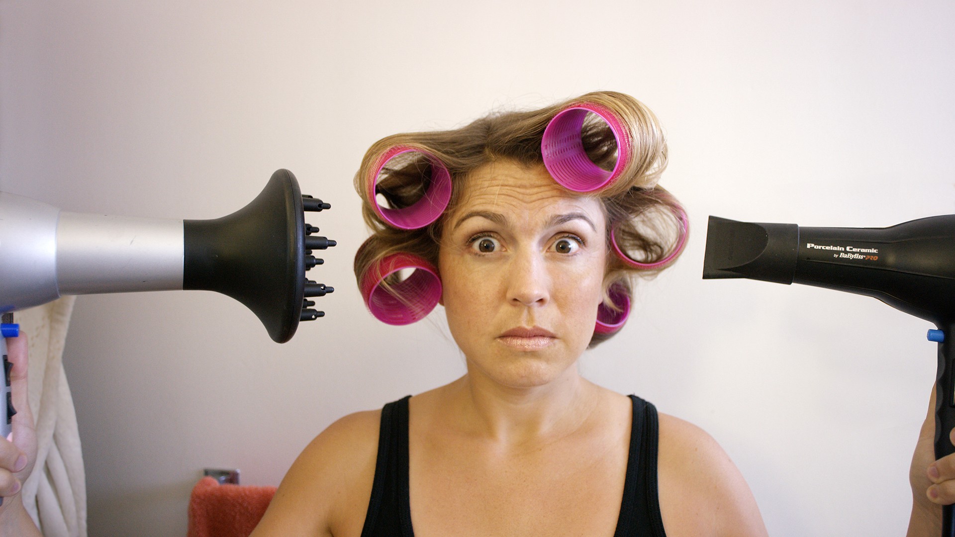 Woman with hair rollers making a surprised face between two blow dryers, from the comedic short Selfie: It's Harder Than You Think