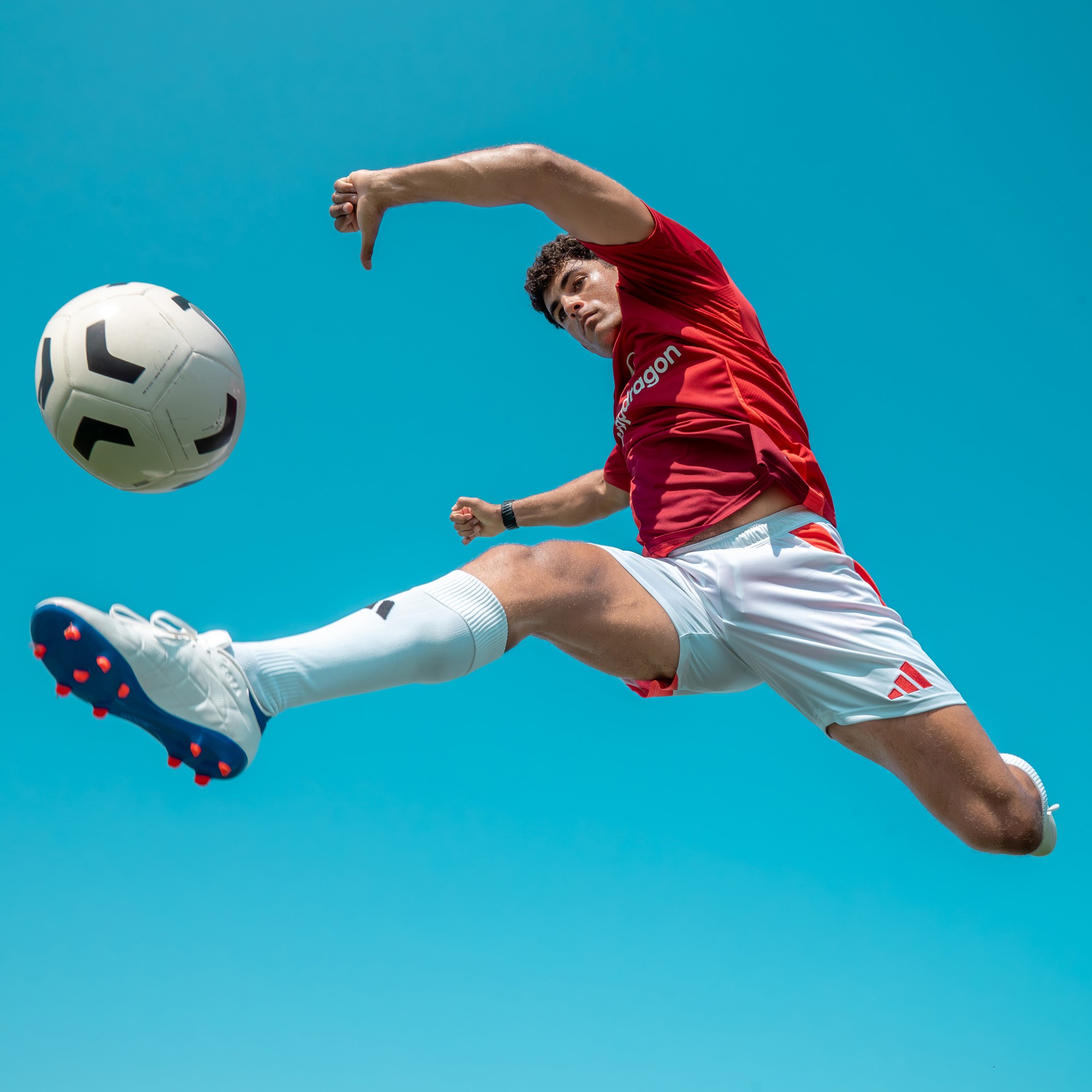 A footballer in a red kit leaping mid-air to strike a ball, against a bright sky.