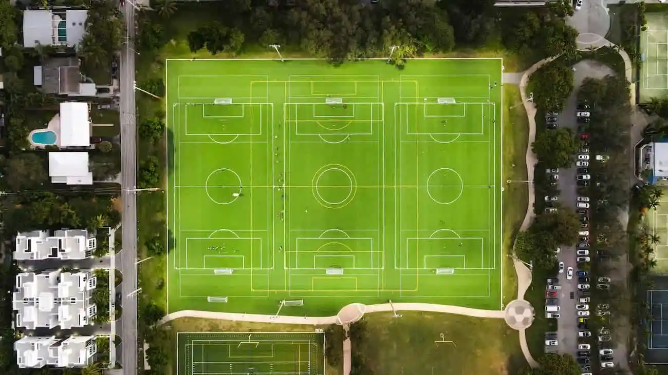 An aerial view captures a meticulously organized sports complex showcasing multiple football fields marked with vibrant white and yellow lines on lush green turf. Each field is for another tournament mode on CoachingArea. The neatly arranged pitches of varying sizes cater to different age groups and skill levels, providing a versatile space for tournaments and coaching sessions. The strategic layout ensures maximum utilization of the space, while a nearby car park, filled with vehicles, indicates the popularity and active use of the facility. The adjacent residential area with a clear boundary line highlights the community's access to recreational sports.