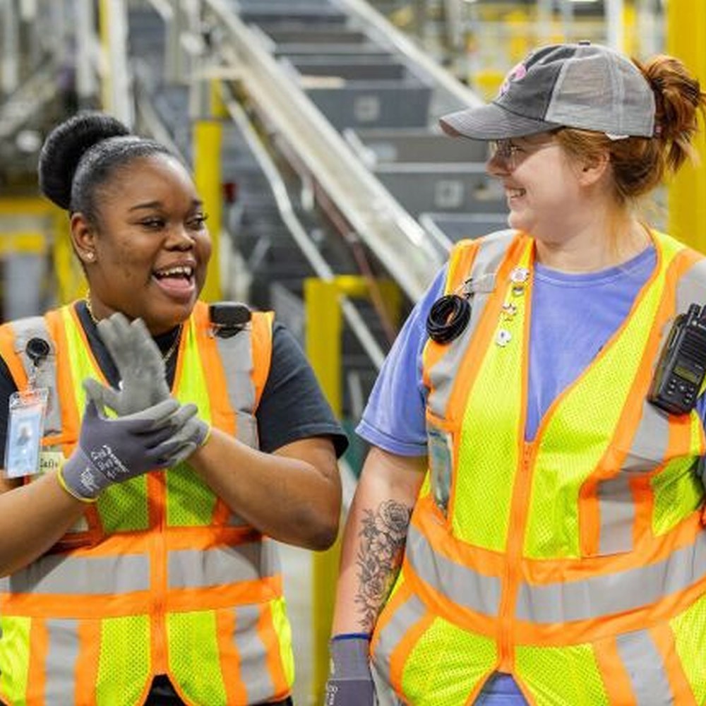 Two women wearing yellow safety vests are conversing in sign language in an Amazon warehouse.