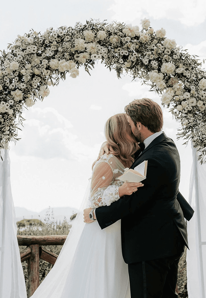 A bride in a white dress and a groom in a black suit hold hands, surrounded by greenery in a romantic setting.