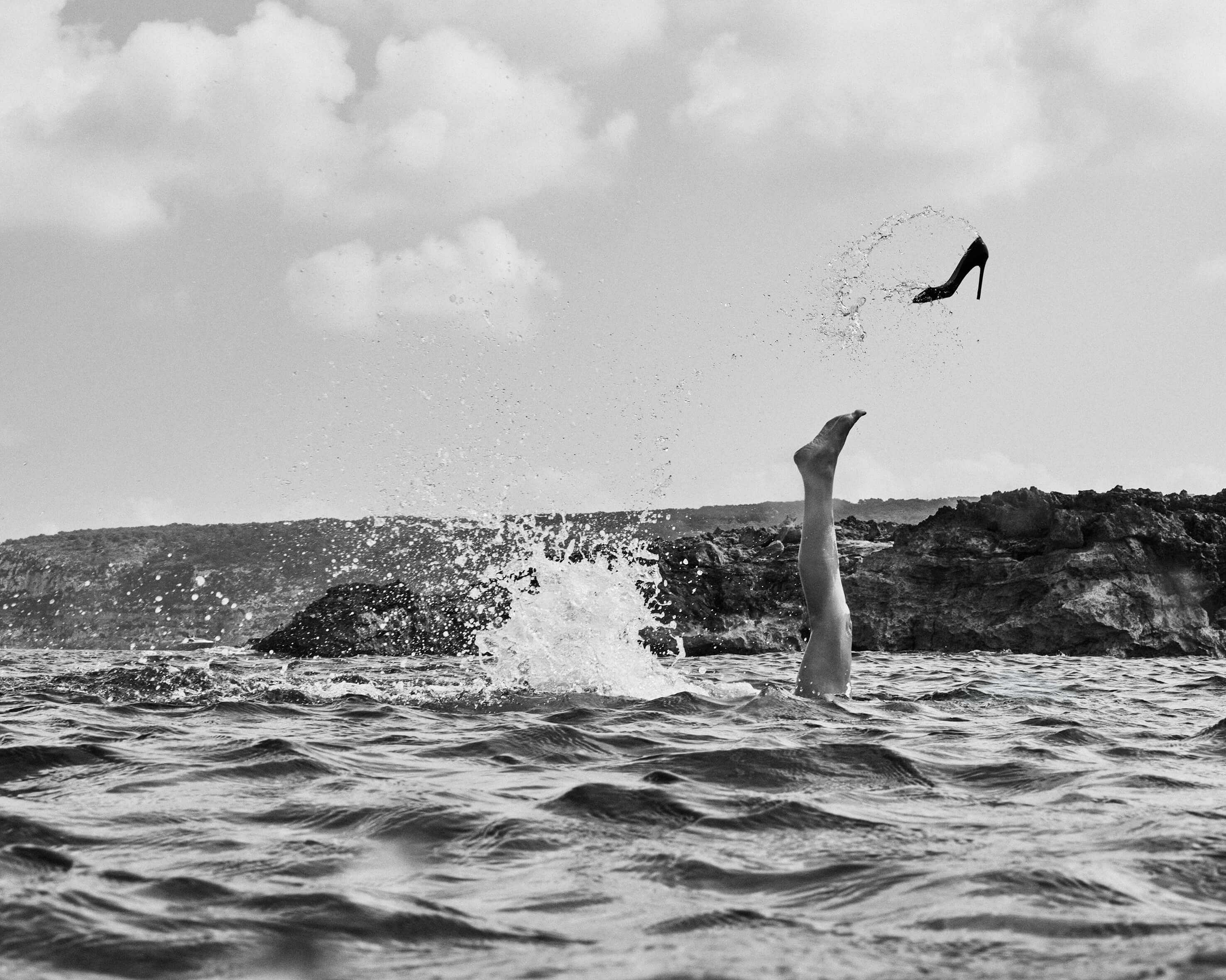 Black and white photograph by José Castellar Carayol: a woman's leg emerges vertically from the sea, throwing a black stiletto heel into the air against a Mediterranean sky and rocky cliffs.