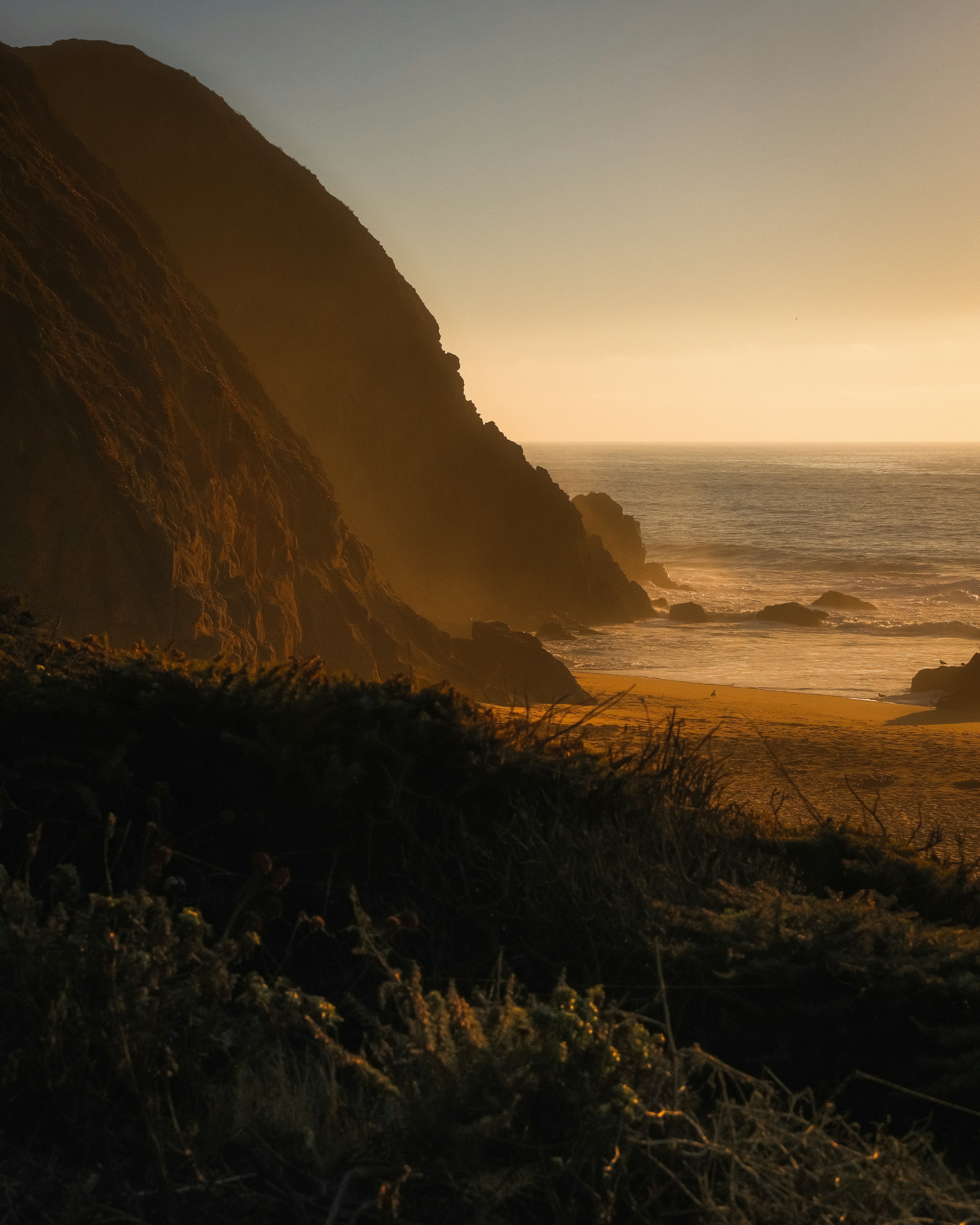 Golden hour illuminates a rugged coastline with ocean waves.