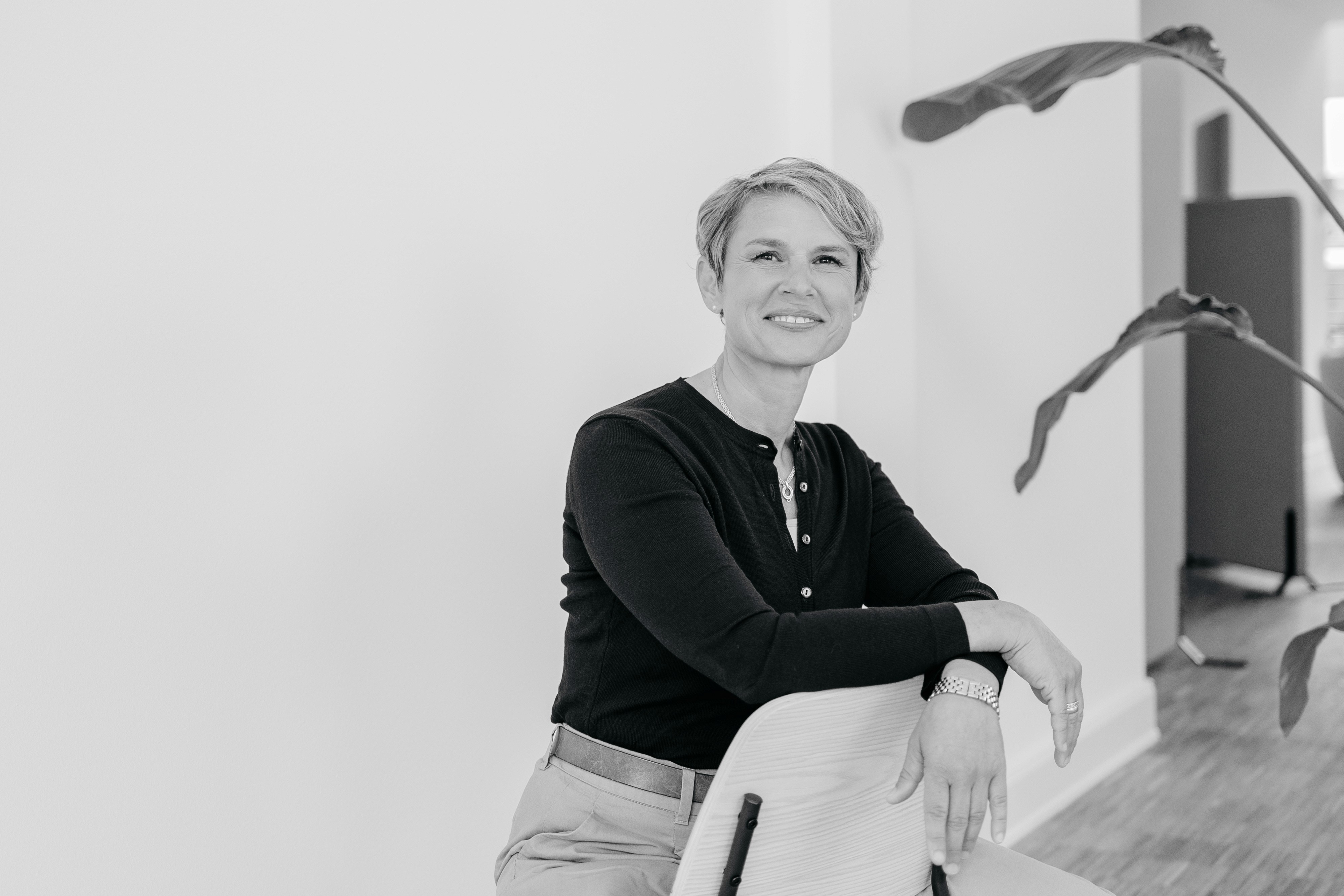 Nathalie Bourgeon sitzt entspannt auf einem Stuhl und lächelt selbstbewusst in einem hellen, minimalistischen Raum
