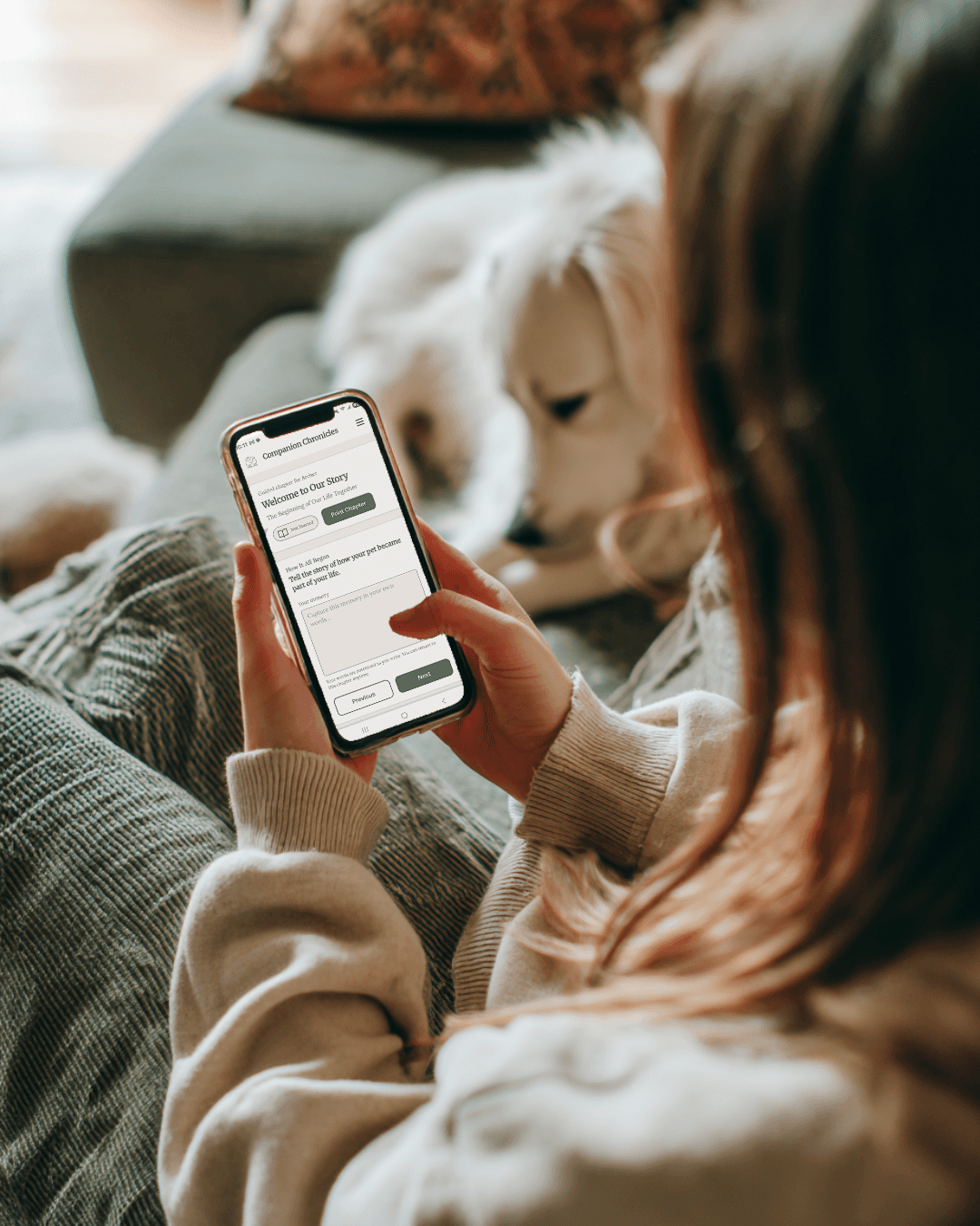 Woman sitting on a couch using a pet memory journal app on her laptop beside her dog.