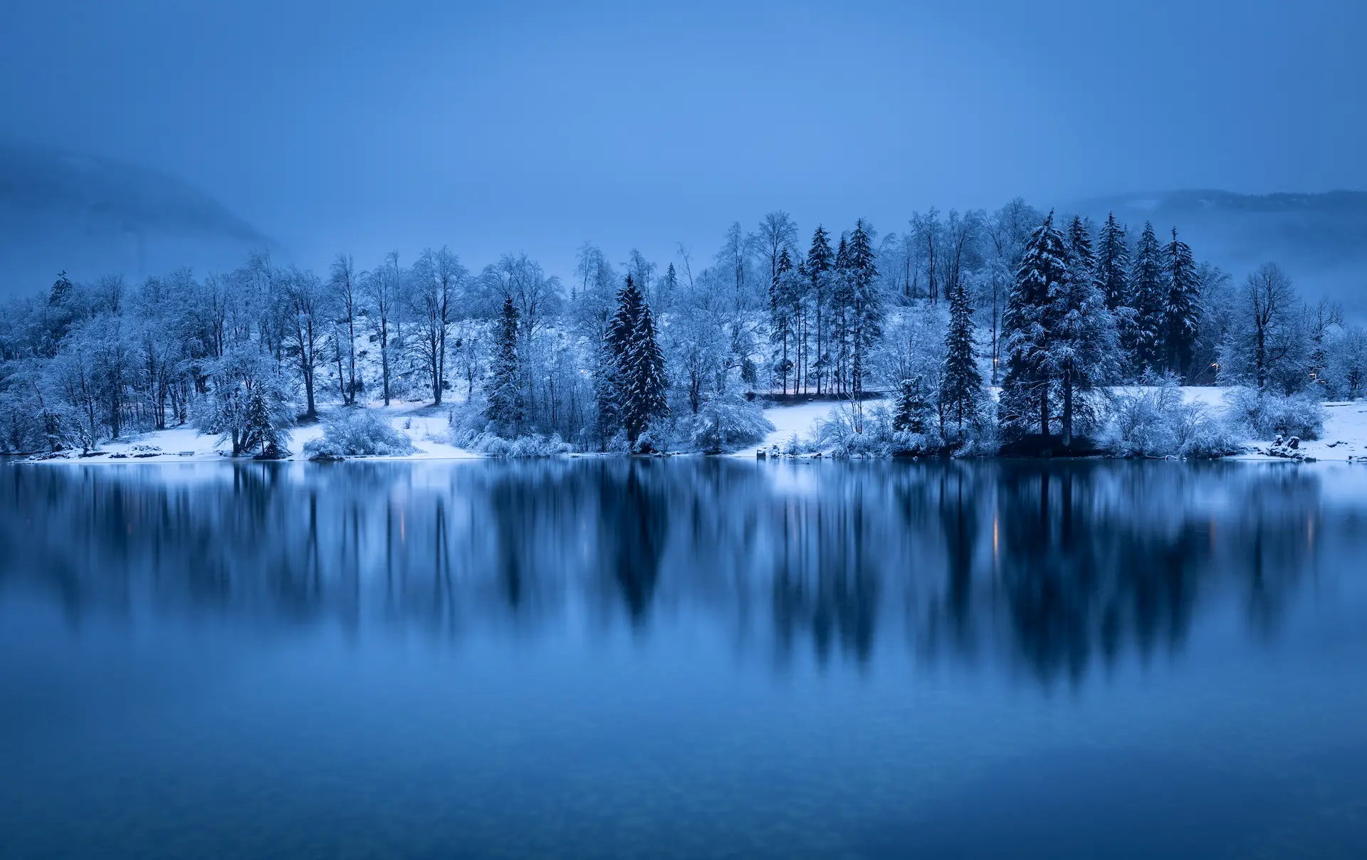 A wide winter landscape of Sveti Tomaž, Slovenia, featuring a hilltop church and scattered houses overlooking a valley of frozen trees under a soft dawn sky.