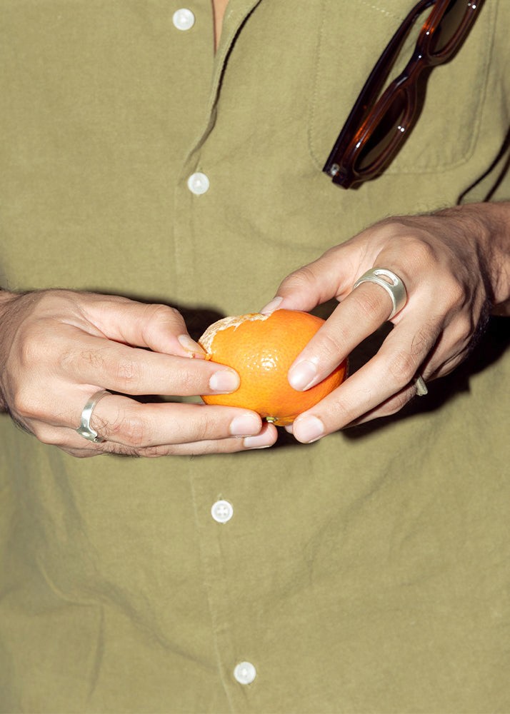 Person holding an orange with a green shirt and sunglasses, wearing silver rings