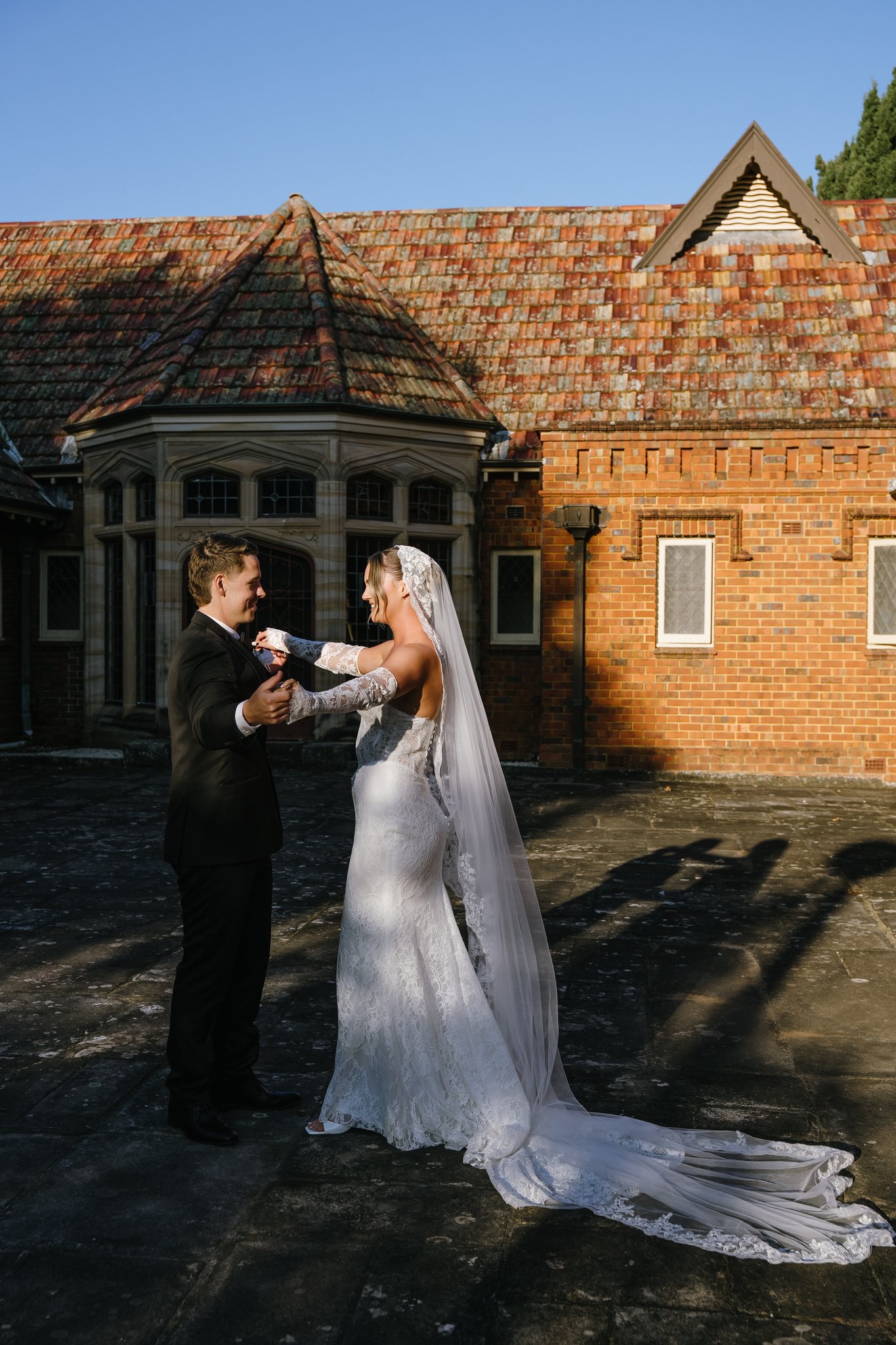 Bride and groom face each other in low golden sunlight casting long shadows across a brick mansion courtyard.