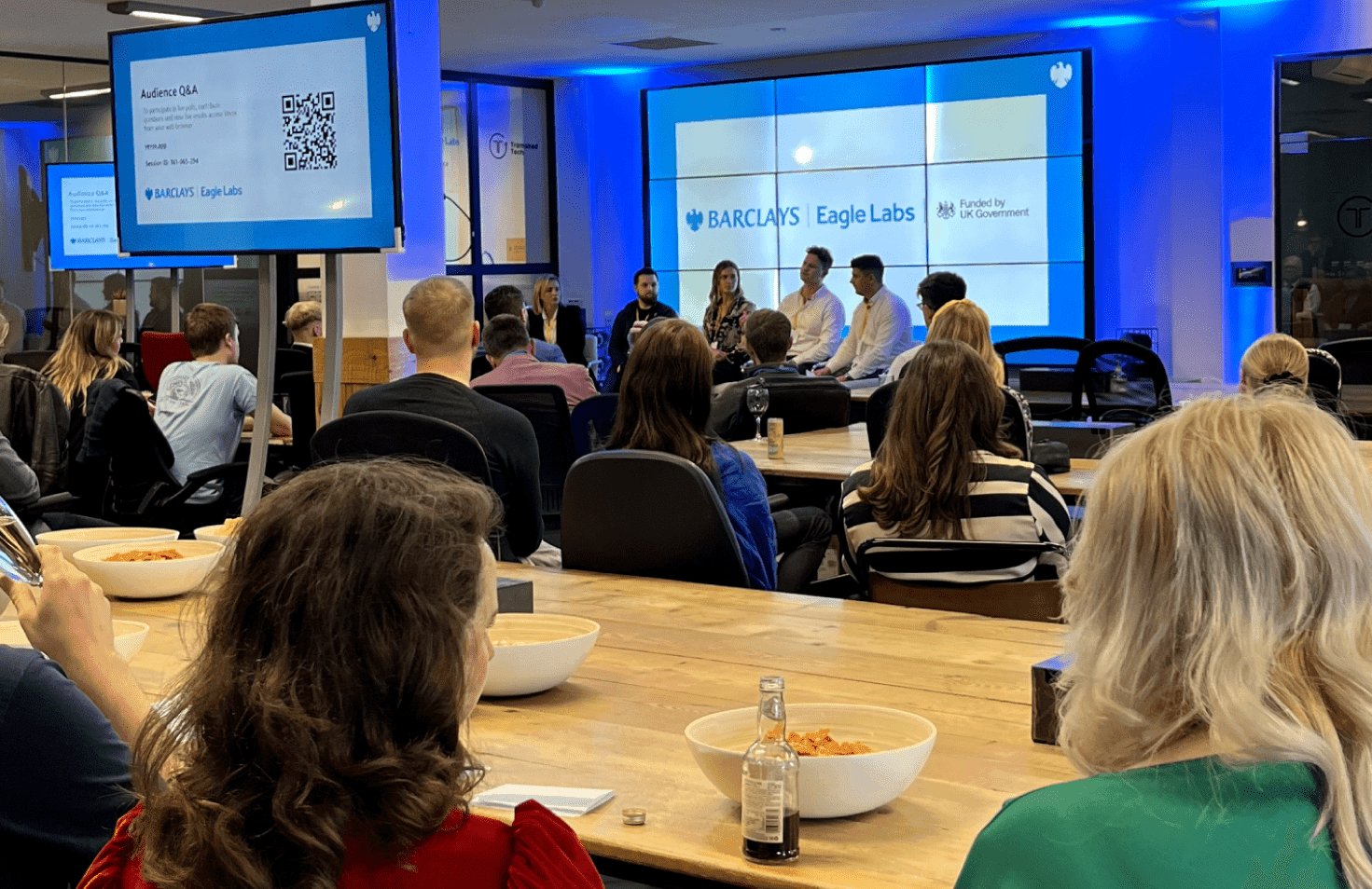 People watching a presentation on laptops in an office. written around the screen and people sitting at tables with chairs, talking to each other while holding papers.