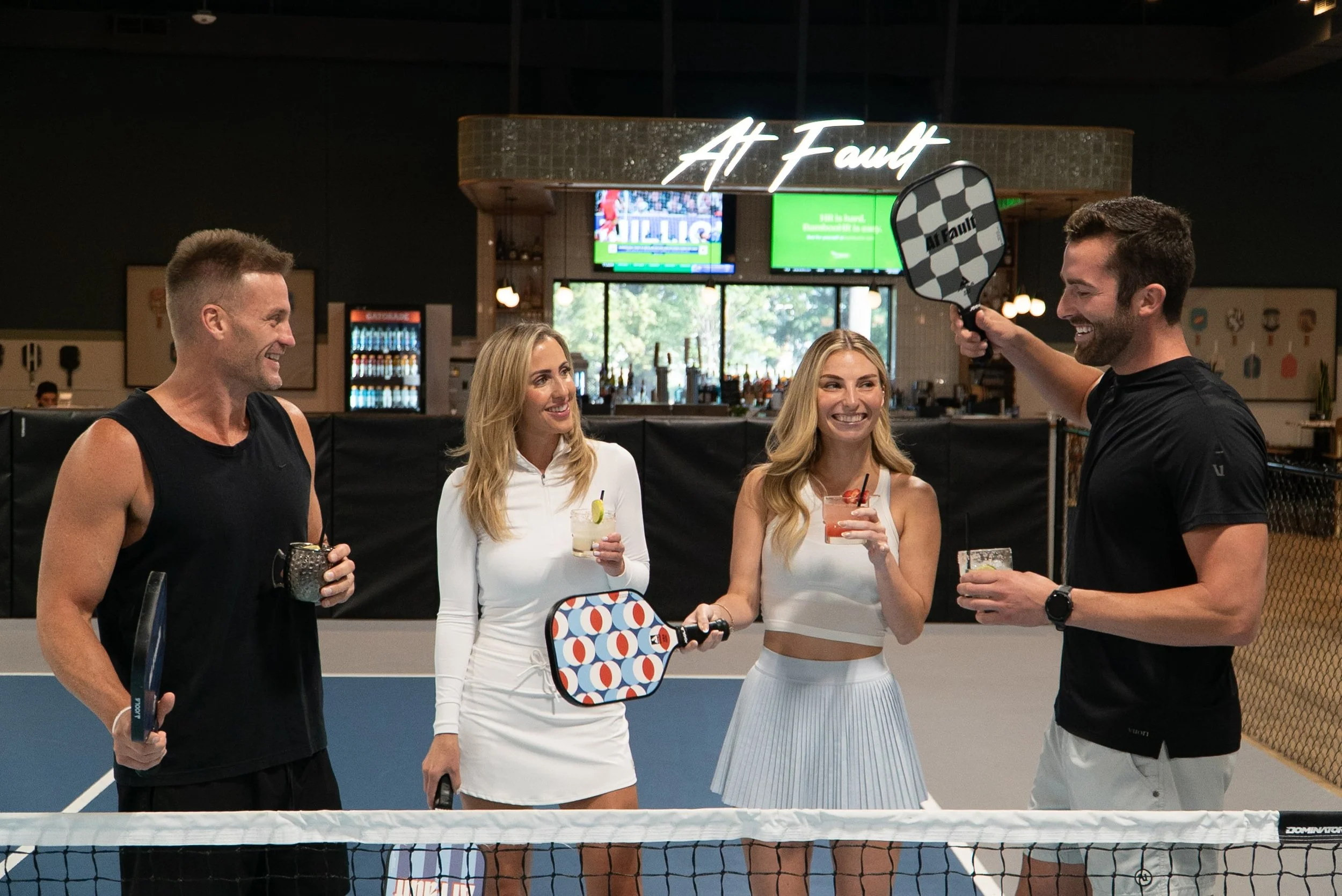Social pickleball play at At Fault in Dallas with players holding paddles and drinks in front of the illuminated bar signage.