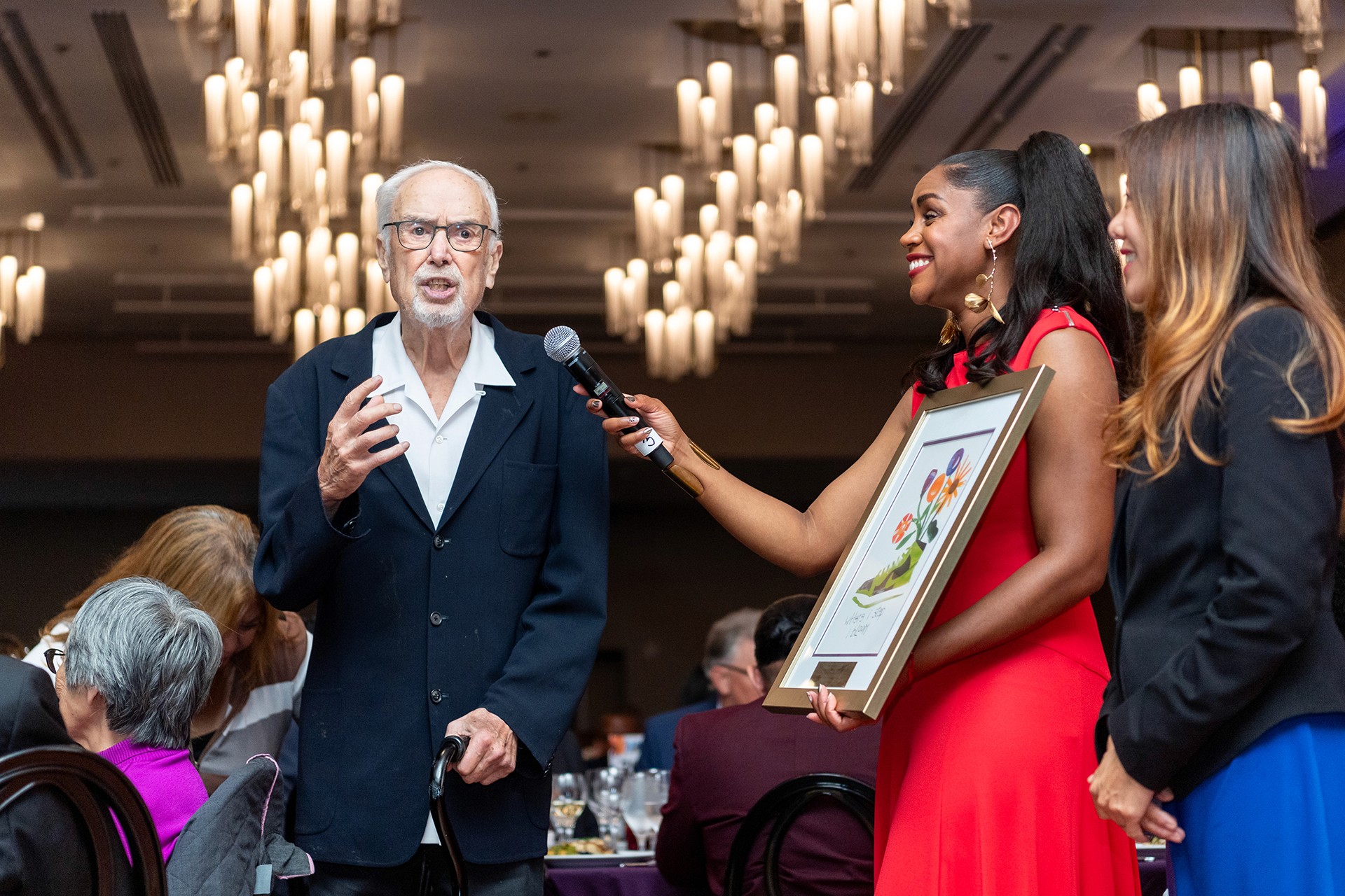 Senator John Burton speaks into a microphone at a gala, an award is presented by Shakirah Simley and California State Treasurer Fiona Ma