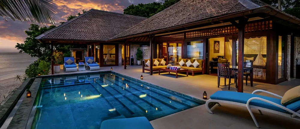 Romantische Dämmerungsszenerie einer luxuriösen Strandvilla mit privatem Infinity-Pool, der blau leuchtet, und einer beleuchteten Holzterrasse, während im Hintergrund ein Sonnenuntergang über dem Meer zu sehen ist.