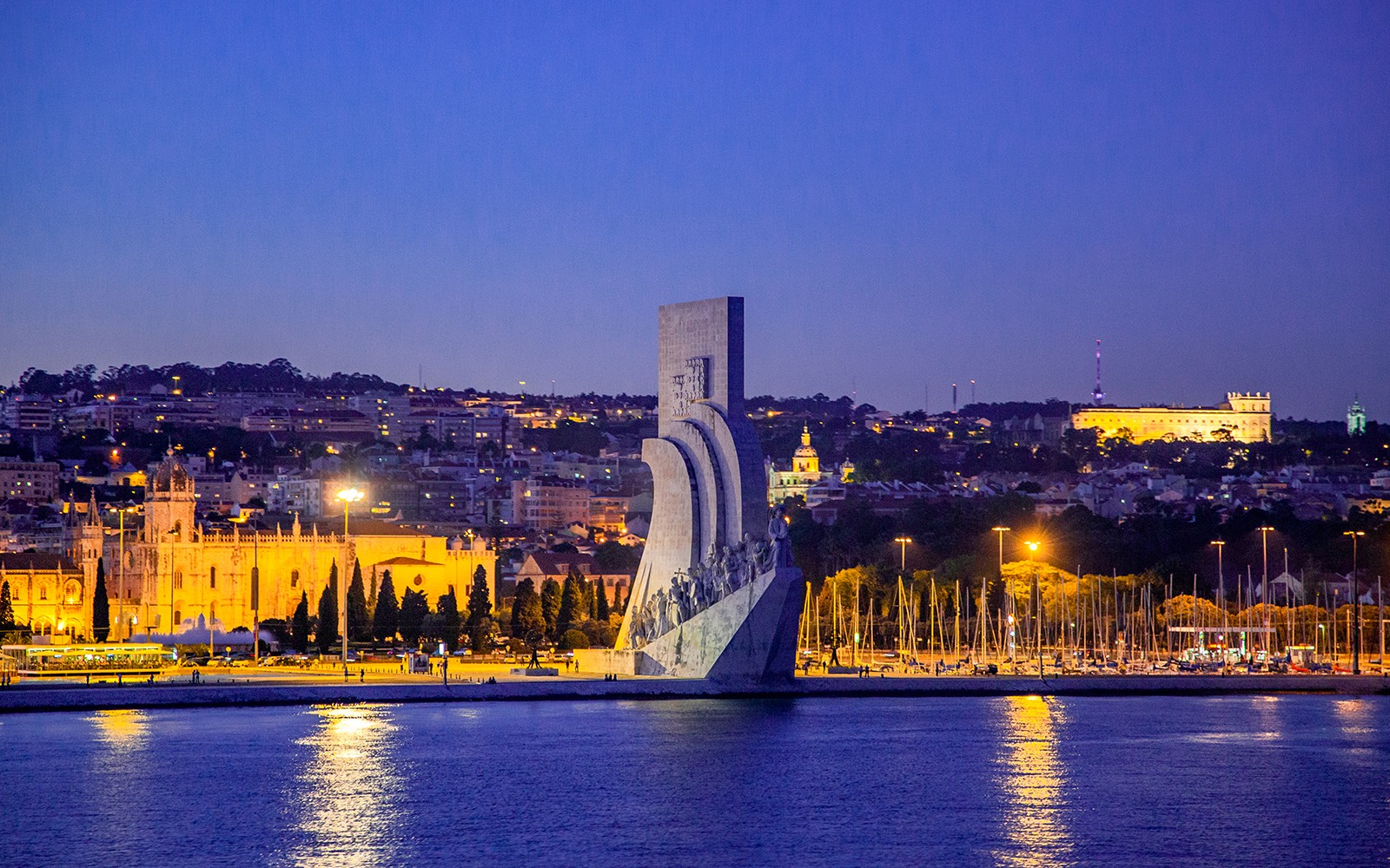 Vista noturna de Lisboa com o Monumento aos Descobrimentos e a paisagem urbana iluminada.