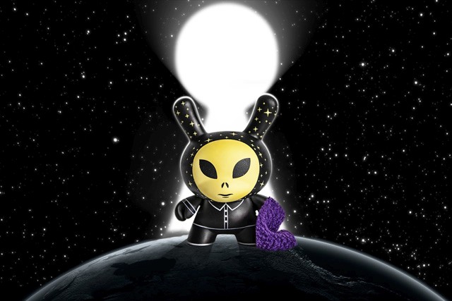 a small alien figurine painted in neon green with the hood with small stars painted with glow in the dark green, and behind it is a galaxy backdrop with small stars all around
