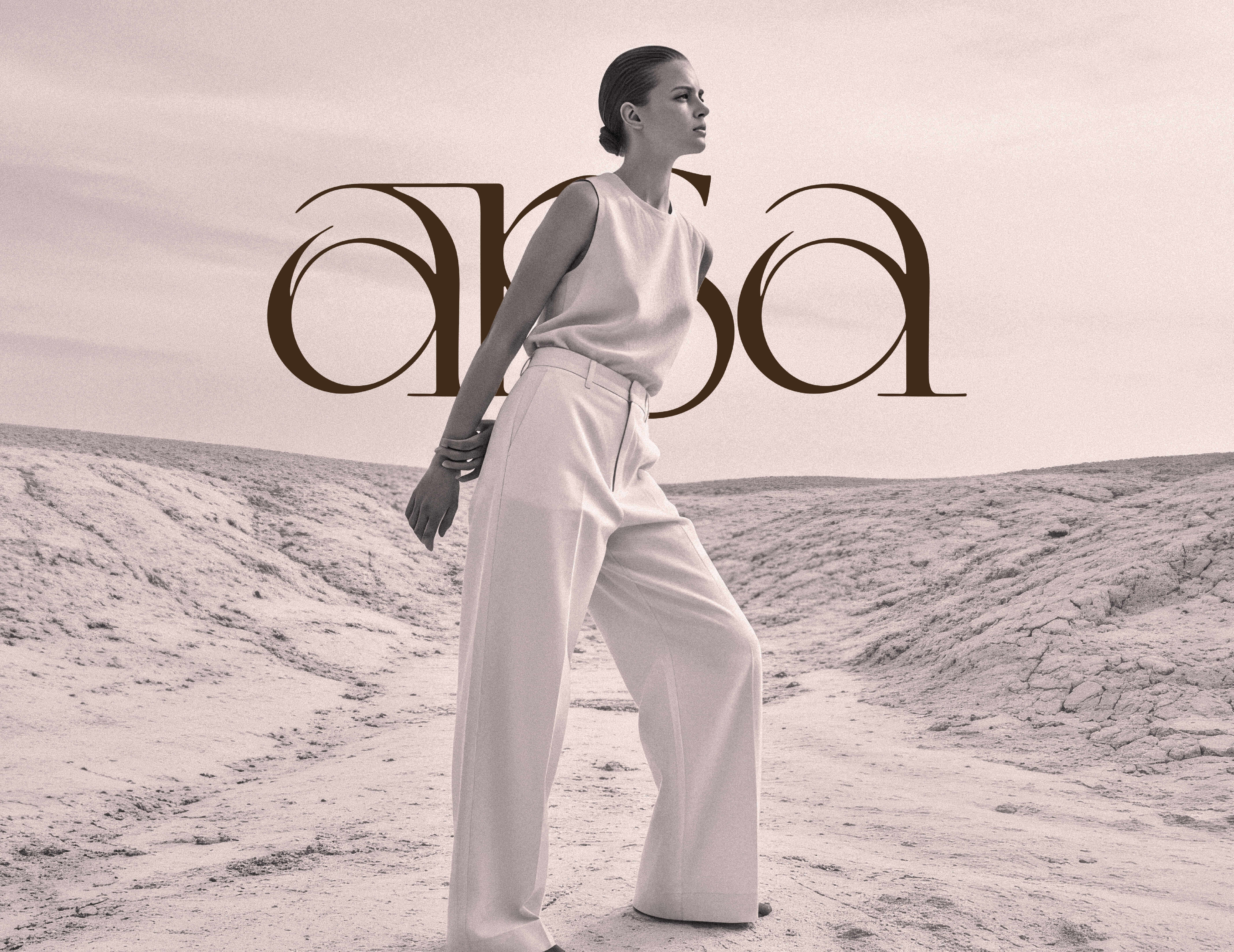 A stylish model with a sleek bun poses in a vast, arid landscape, wearing a light-colored, sleeveless top and loose-fitting, high-waisted trousers. The scene is presented in a muted palette, evoking a soft, vintage aesthetic. Prominently featured in the foreground is the word arisa in bold, artistic type, enhancing the fashion-forward vibe of the image. The backdrop consists of cracked earth and subtle hills, contributing to a chic, minimalist atmosphere.