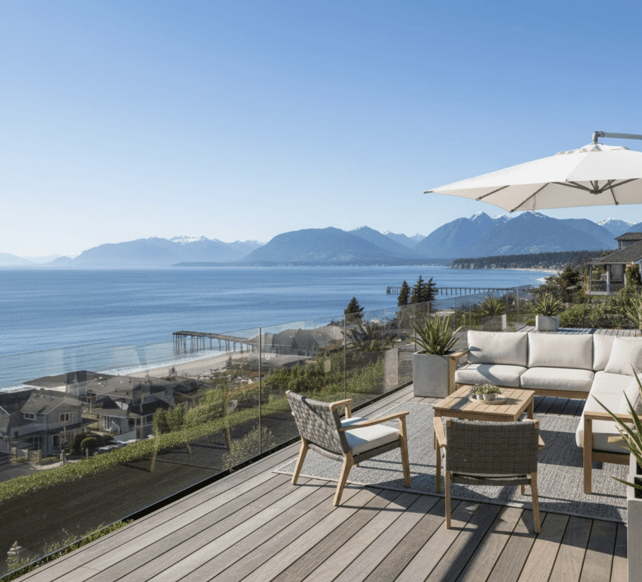 Wide angle shot of White Rock residential deck with frameless glass railings, panoramic ocean views, mountains in distance, modern outdoor furniture, sunny day, showing full sightlines through glass