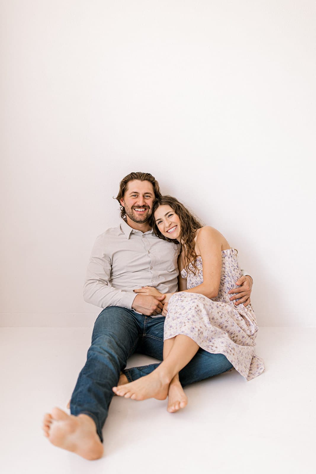 A couple sits closely on the floor, smiling together during a natural light engagement session at Revelator Studio in Shreveport.