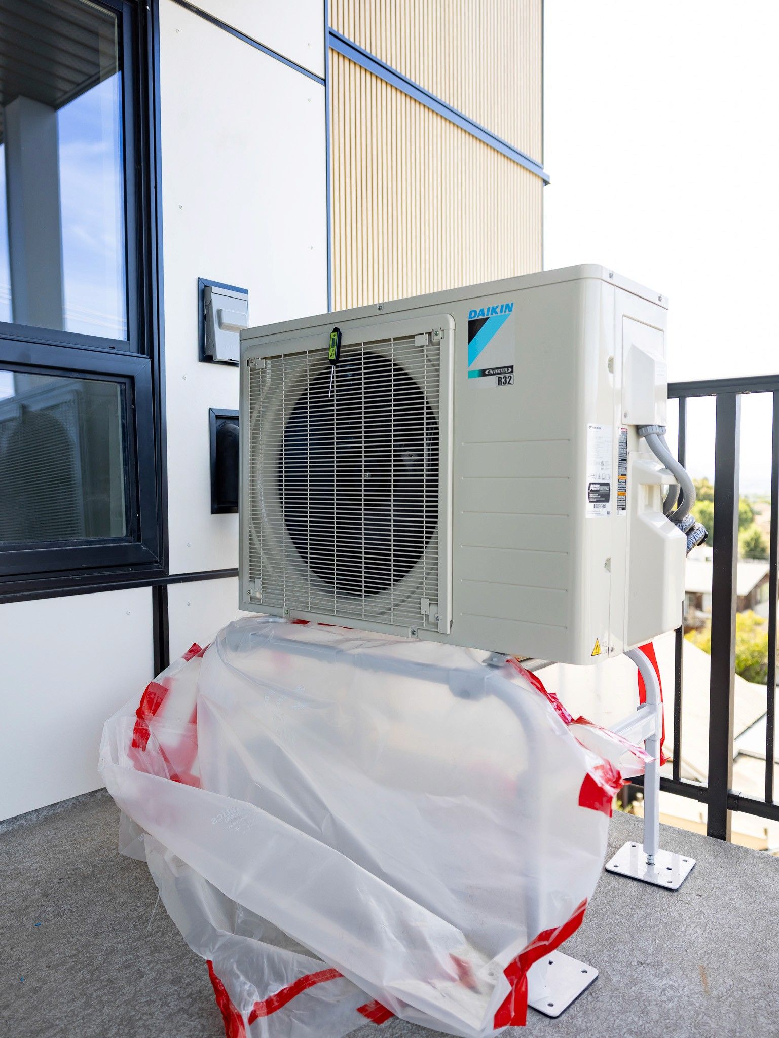 An air conditioning unit installed on a balcony, partially covered with plastic, situated next to a glass door.