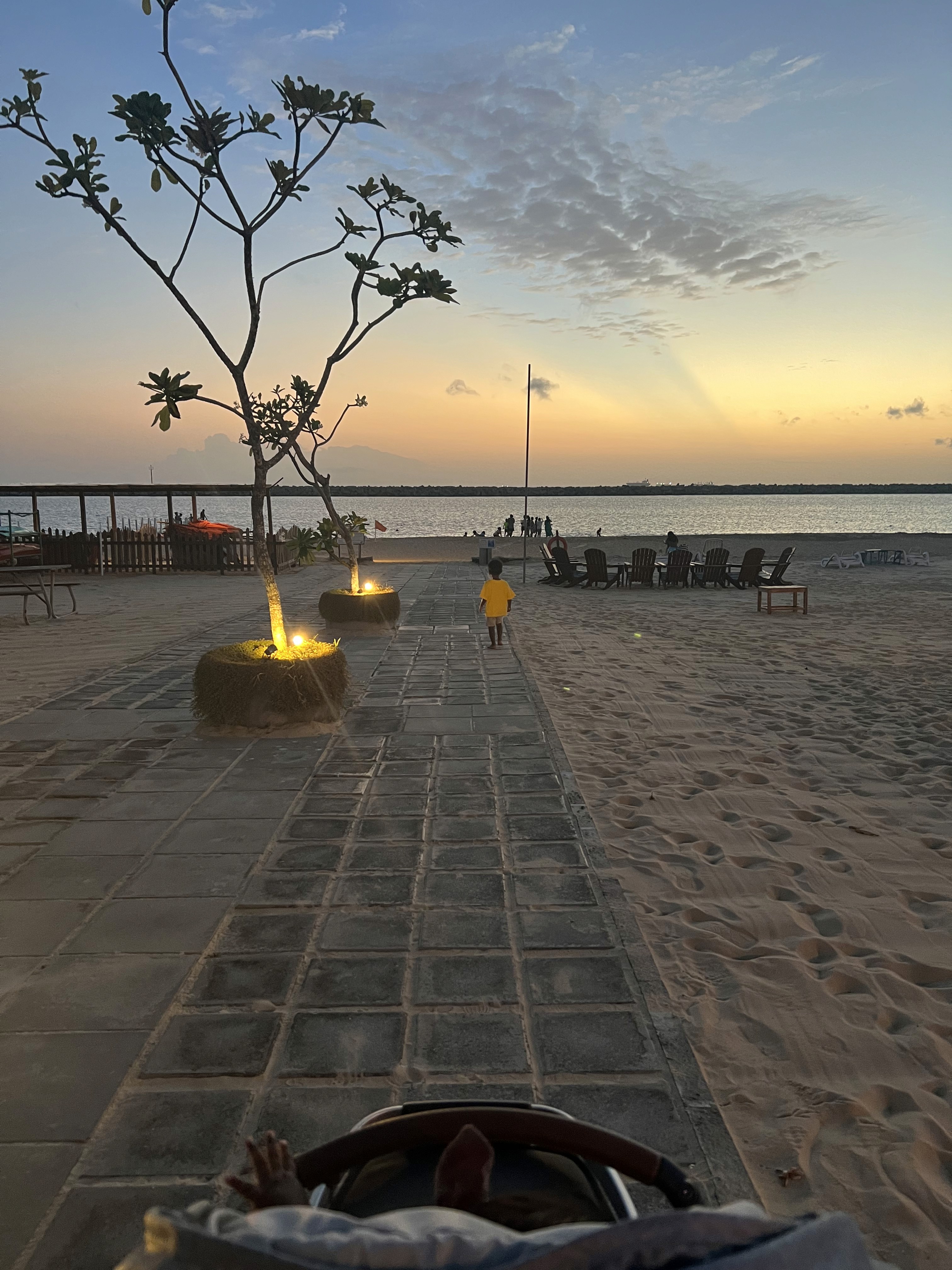 A child in a yellow shirt walks toward the ocean at sunset at Beach Park in Colombo, seen from behind a stroller.