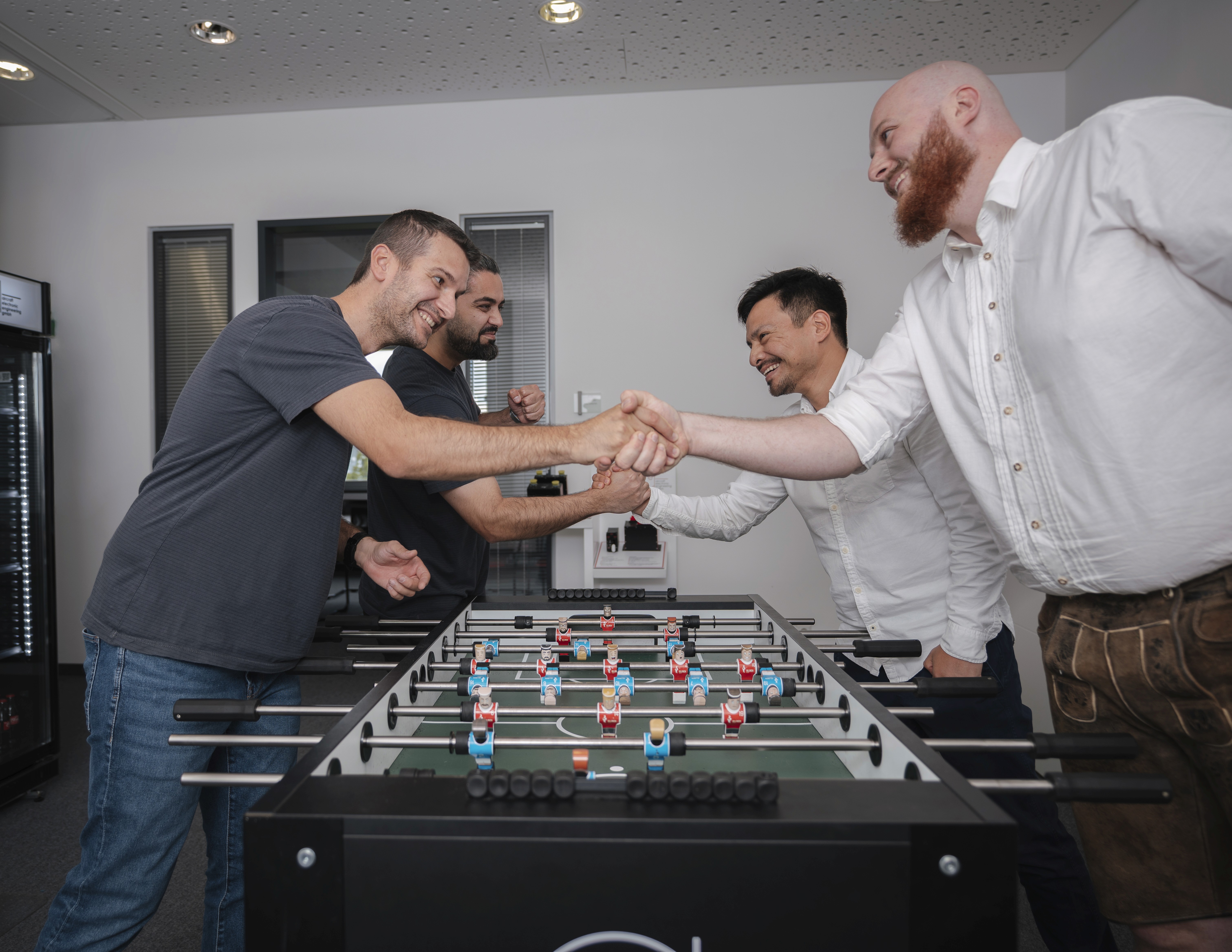 Four men at a foosball table, two on each side, shaking hands across the table in a gesture of sportsmanship.