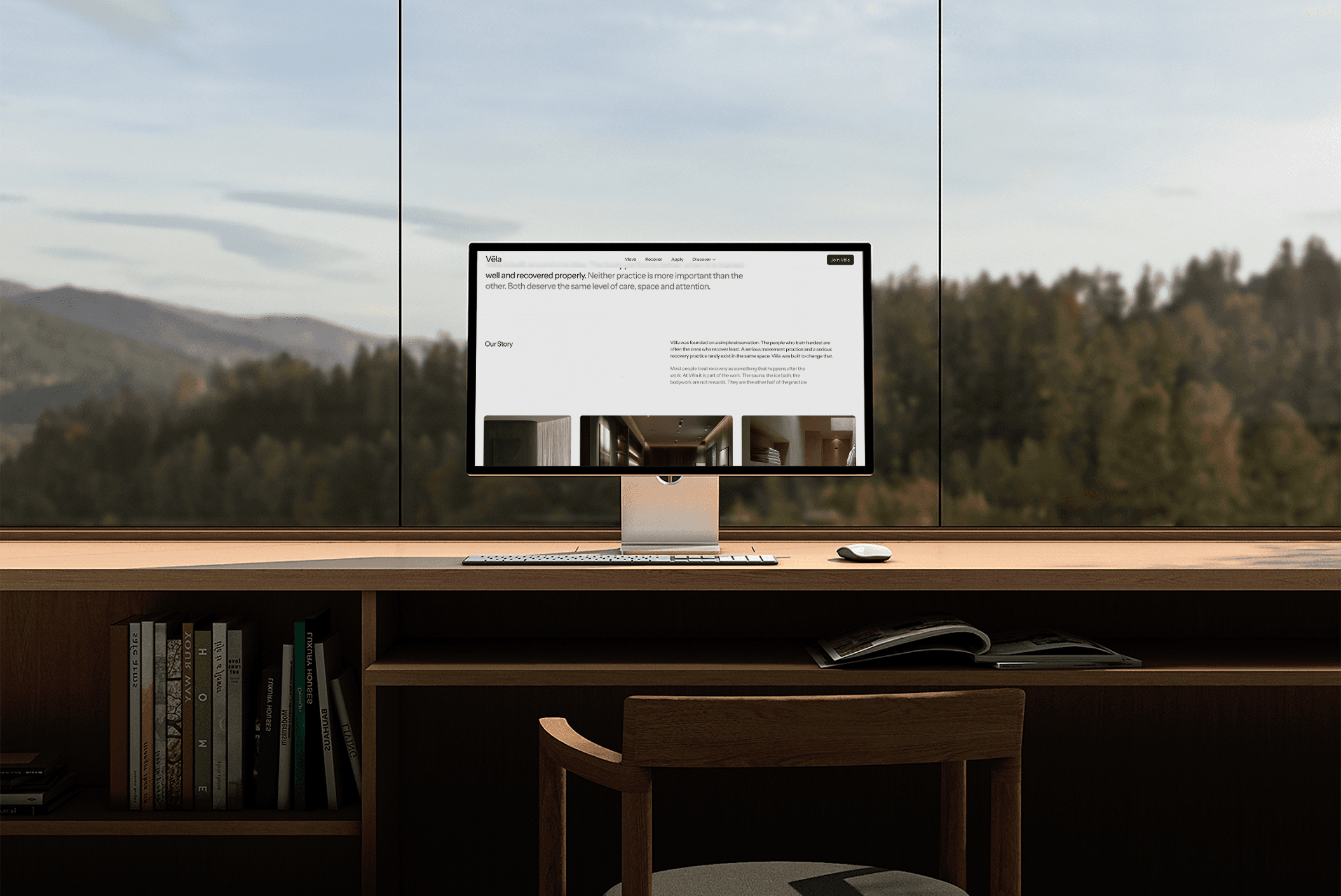 A desktop computer displaying the Véla website, set on a wooden desk against a floor-to-ceiling window overlooking a forested mountain landscape.