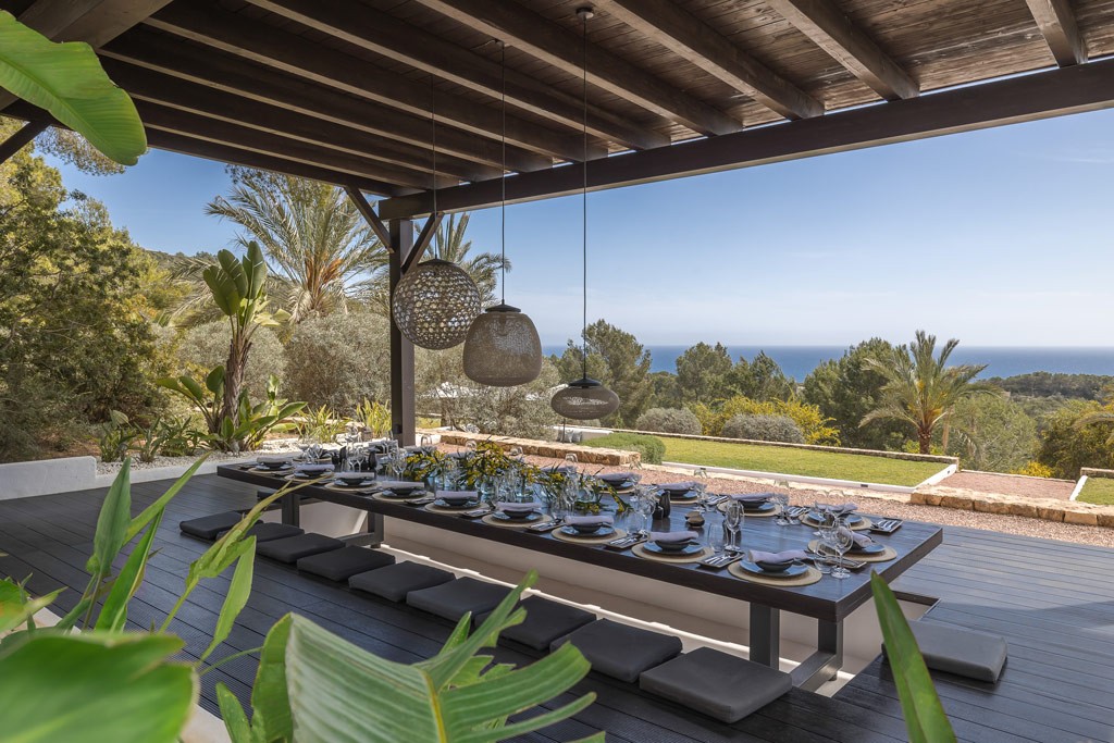 Covered outdoor dining terrace with long table setting overlooking Mediterranean landscape and sea horizon.