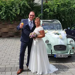 Glückliches Hochzeitsfeedback - Braut Michaela im eleganten Brautkleid Arm in arm mit ihrem Mann vor dem nostalgischen Ente-Hochzeitsauto