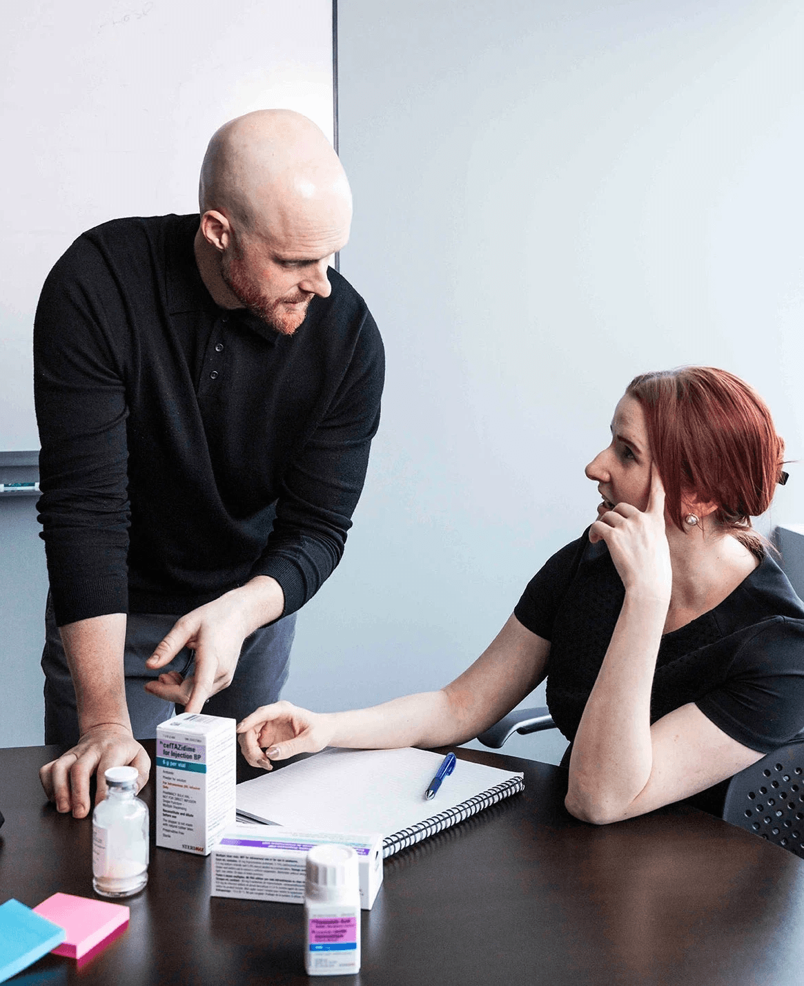 Two colleagues have a discussion at an office table with notes and pill bottles.