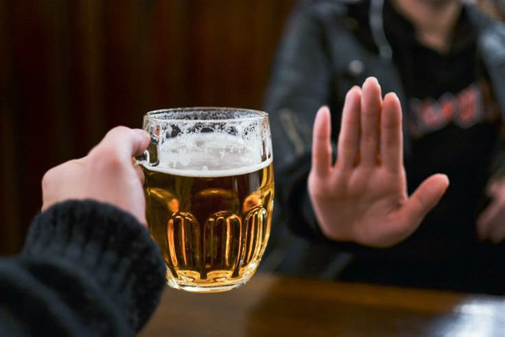 A person extends a hand holding a mug of beer toward another person, who gestures "stop" with an open palm, symbolizing refusal of alcohol.