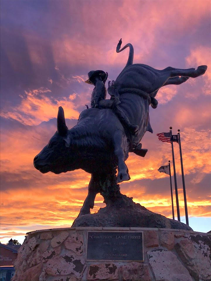 A dramatic silhouette of the bronze "Champion Lane Frost" bull riding monument in Cheyenne, Wyoming, captured against a brilliant sunset with vibrant orange, pink, and purple clouds. The large sculpture of the cowboy riding a bucking bull is positioned on a stone pedestal on the left, while the American flag and Wyoming state flag fly from poles on the right.