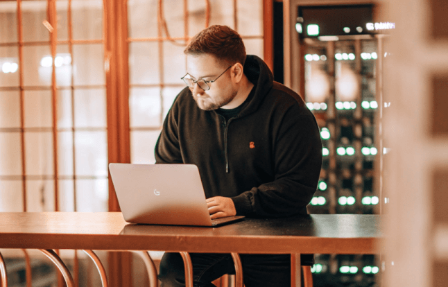 Man in glasses, wearing a black hoodie, working on a laptop at a wooden table in a well-lit room.