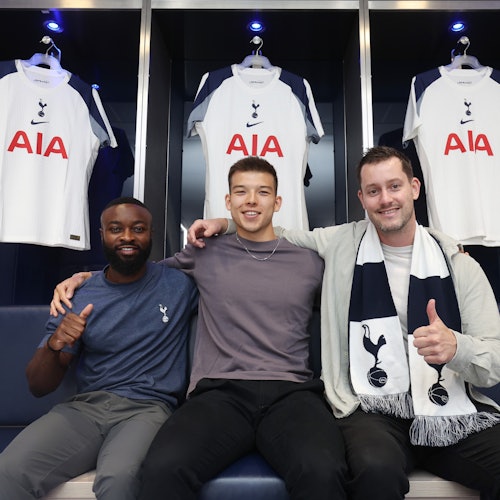 Three men sit on a bench in a locker room, each giving a thumbs-up, with Tottenham Hotspur football jerseys hanging behind them.