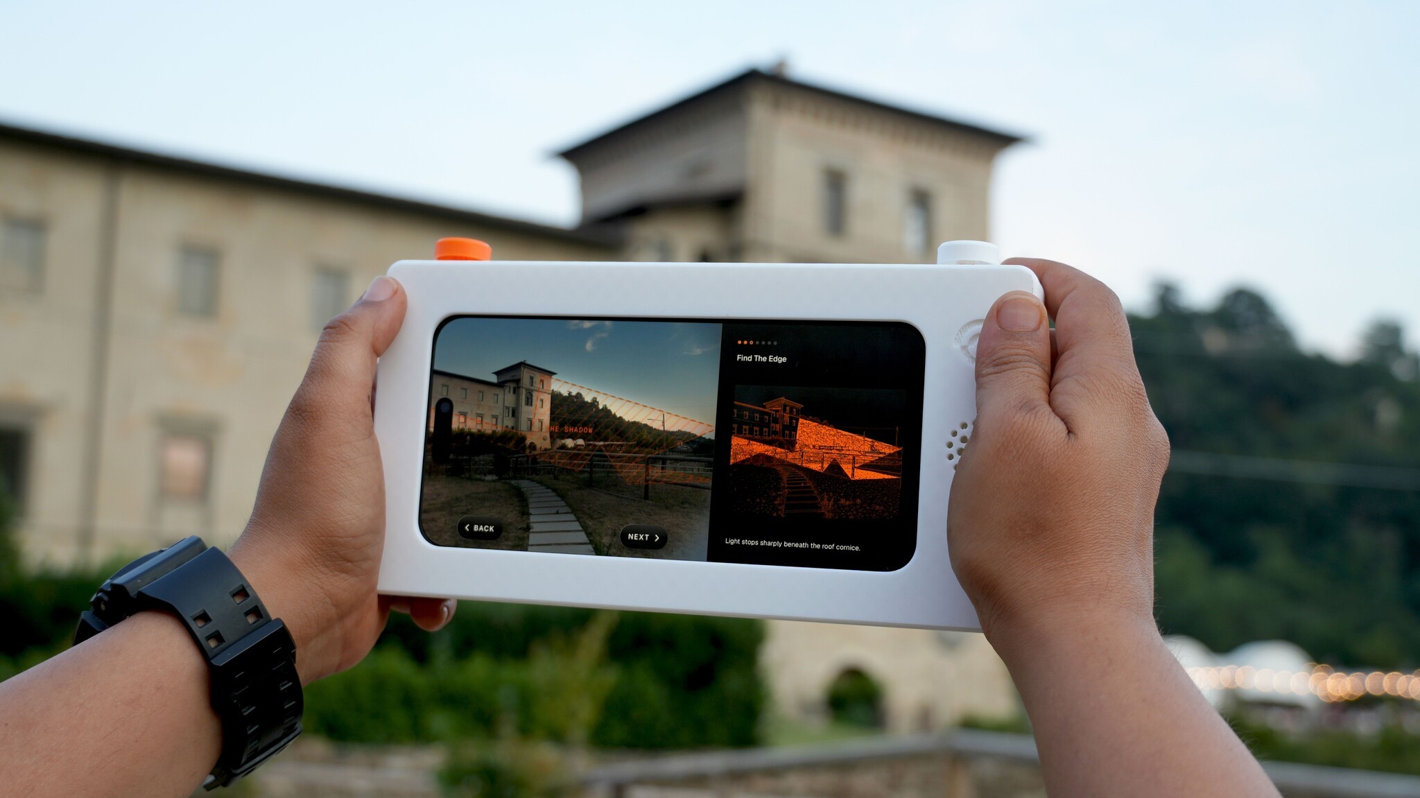 Two hands holding the Attune device up to a hillside building, its screen showing photos