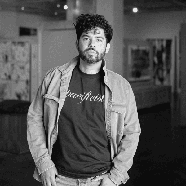 A black and white photo of Freddy Janka with his hands in his pockets. There is framed art in the background.